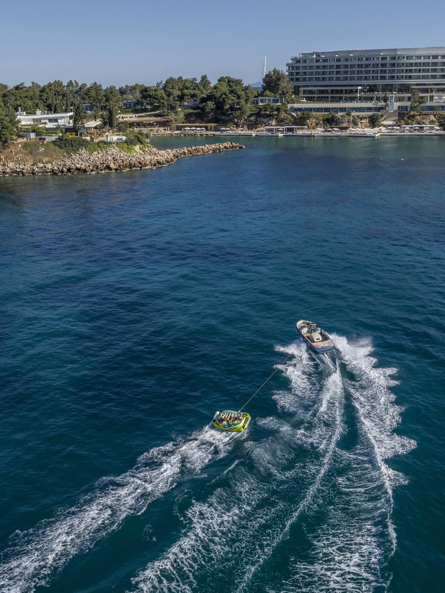 Speedboat pulls raft of people on blue Aegean ocean with building and shoreline in background
