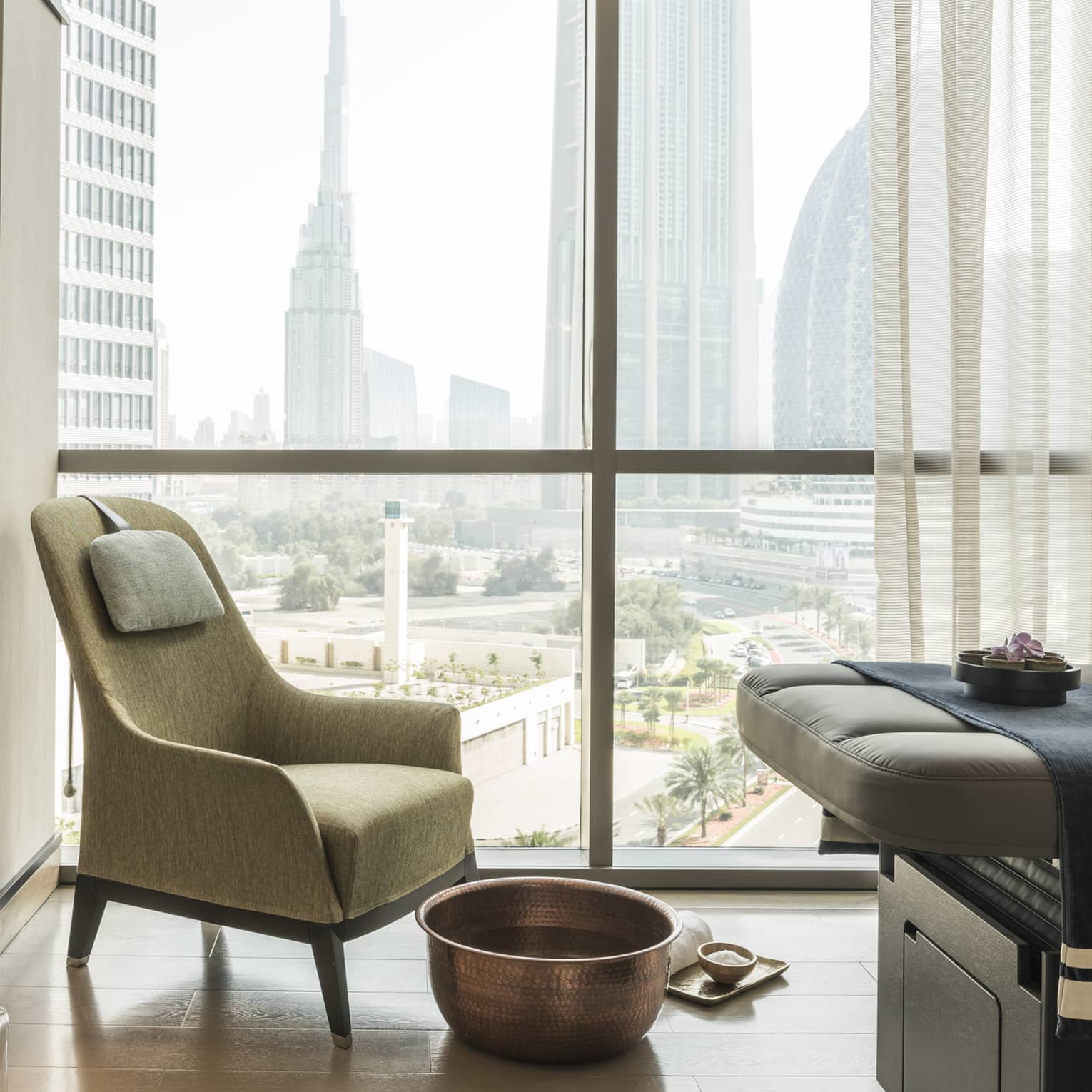 Armchair in hotel room corner by sunny window, large brass bowl foot bath, rolled white towel on floor