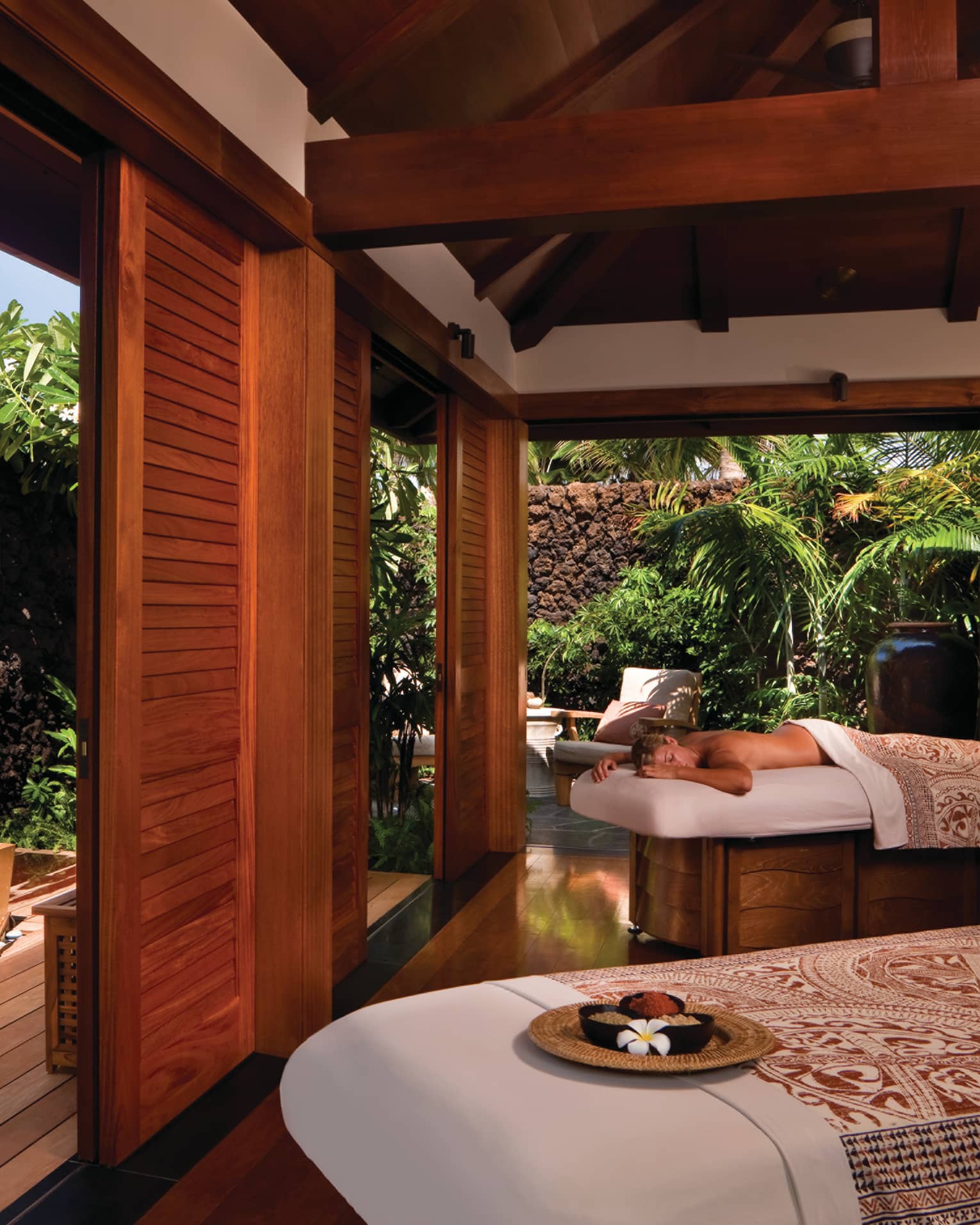 Woman lies on massage table near man soaking in spa tub in sunny Couples suite 