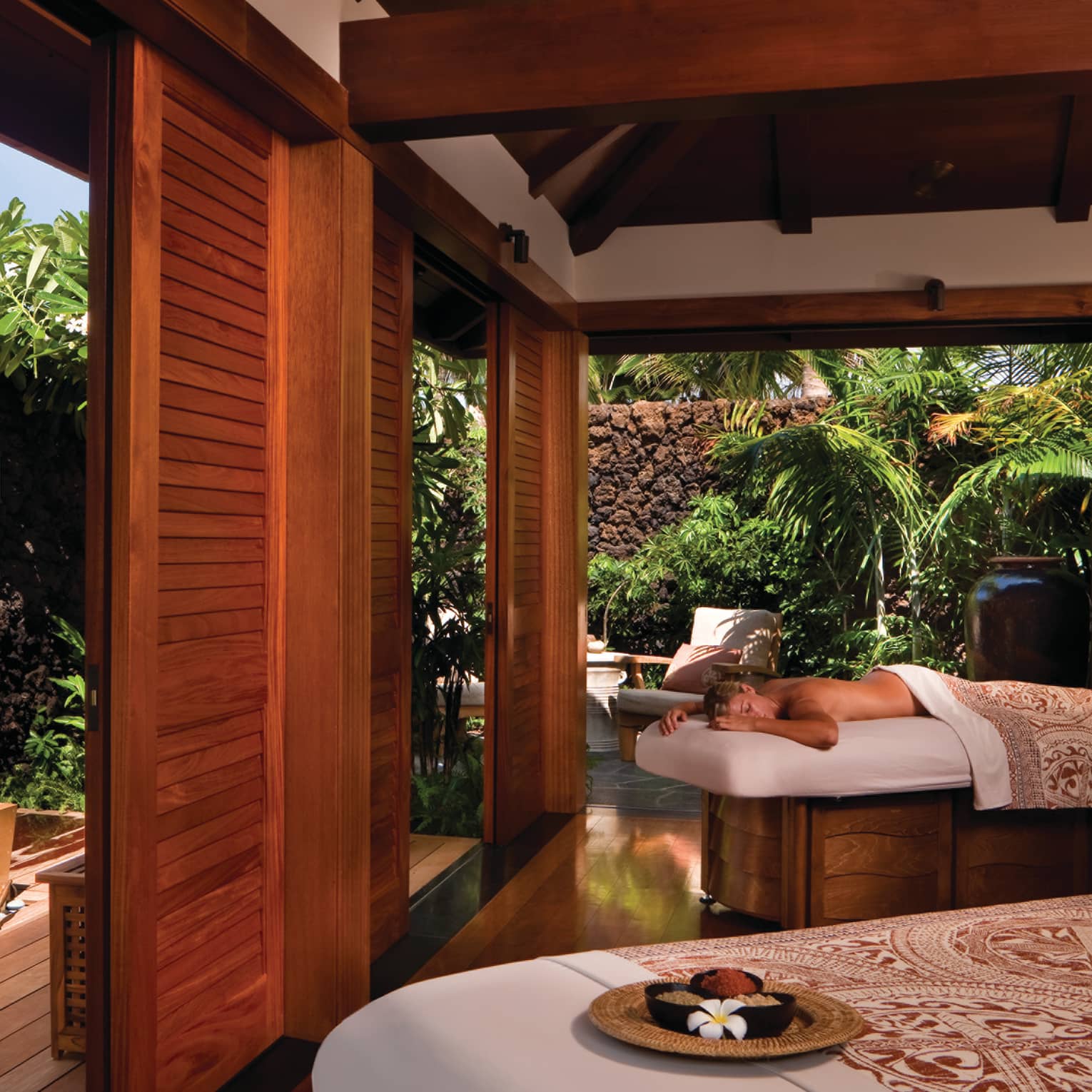 Woman lies on massage table near man soaking in spa tub in sunny Couples suite 