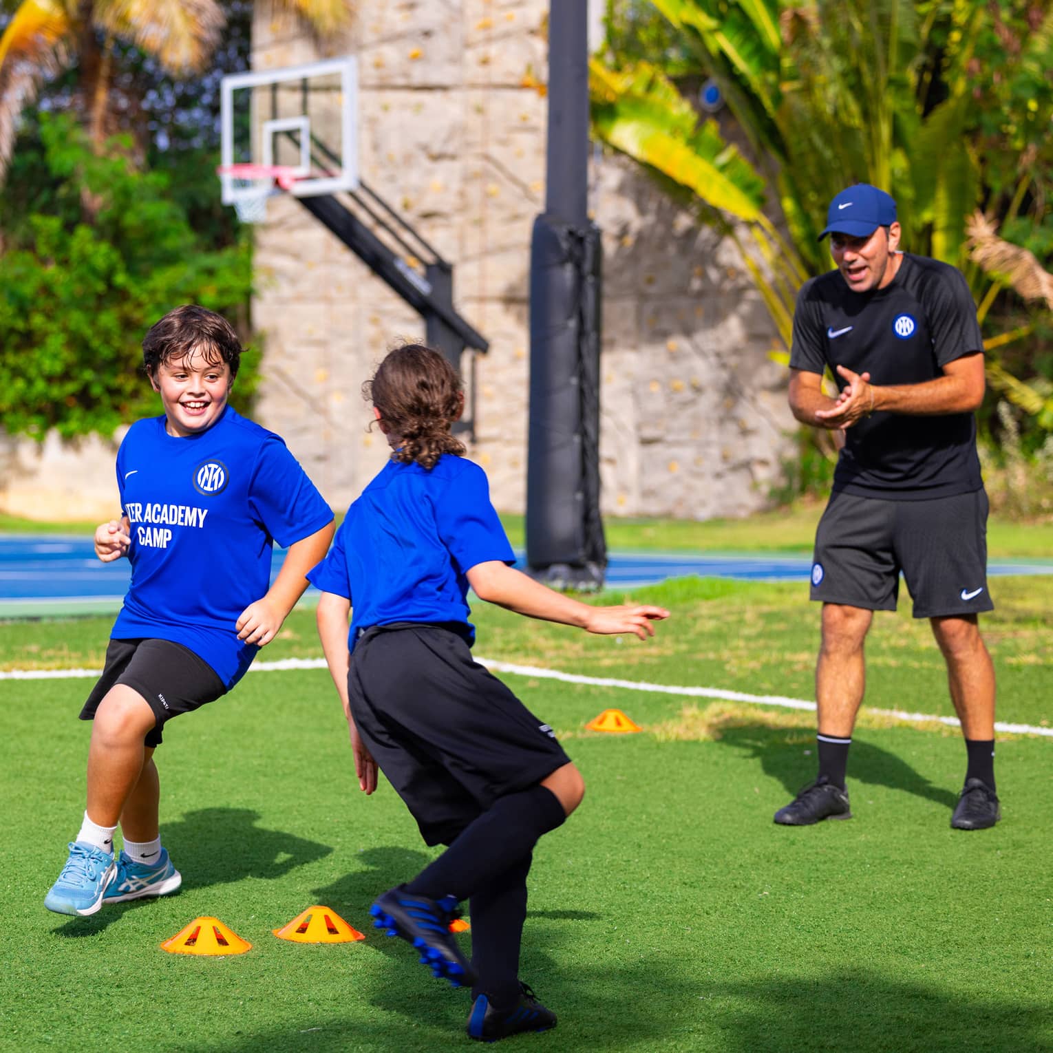 Soccer coach talking to two young guests exercising outside with a basketball court in the background.