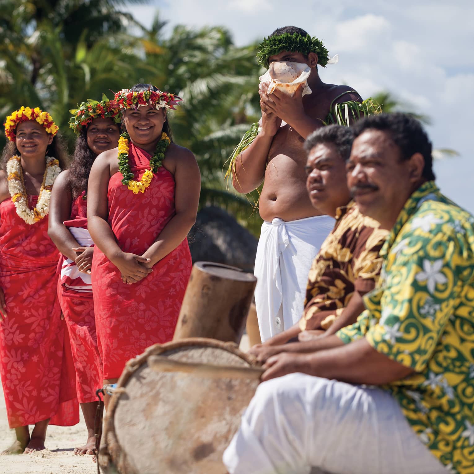 Group of people in traditional attire with leis and a conch shell, standing on a beach