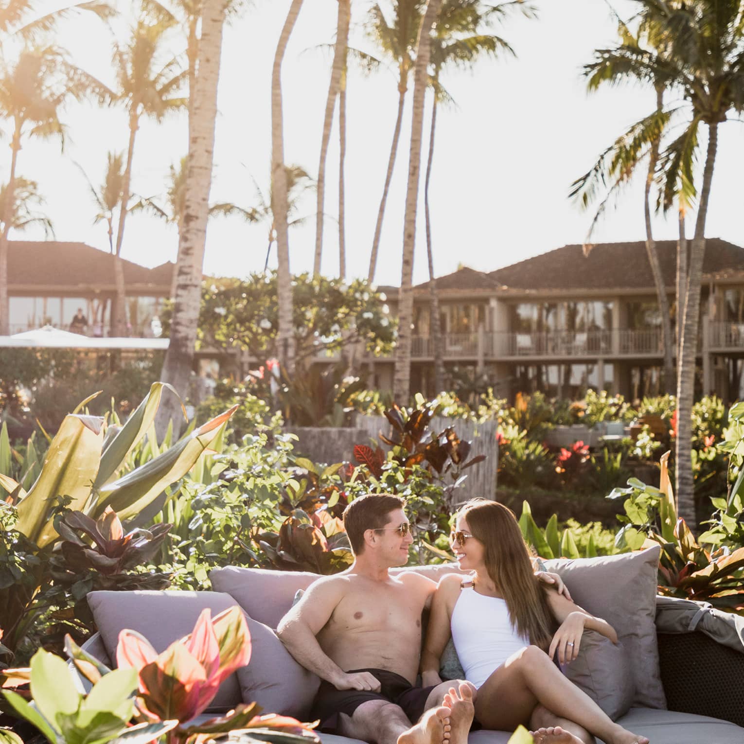 A couple lounges on a round cushioned sofa-bed nestled within lush, tropical foliage, as the sun beams through palm trees.