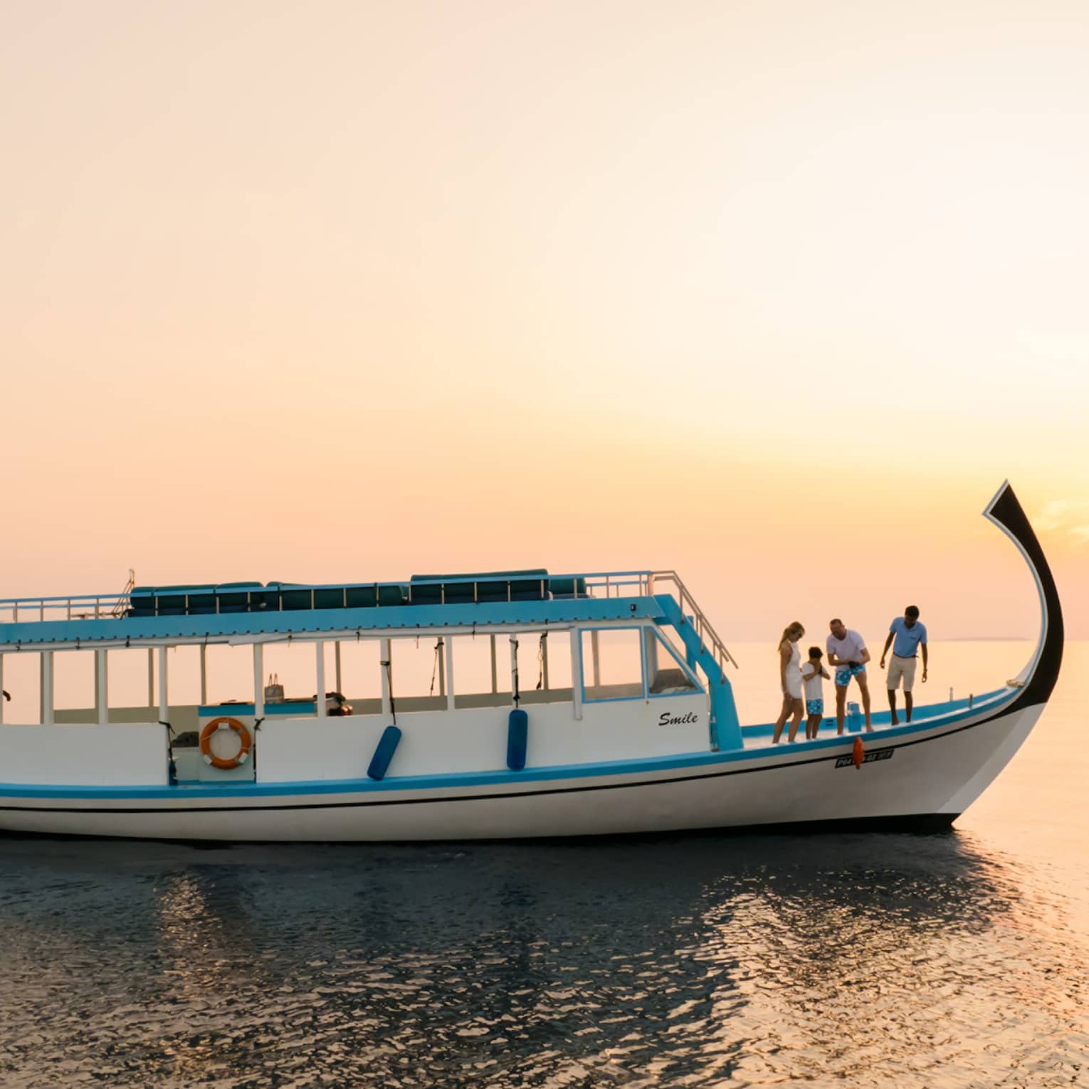 In sunset's golden glow, four people on the prow of a dhoni boat peer over the side as one pulls up a red fish on a line.