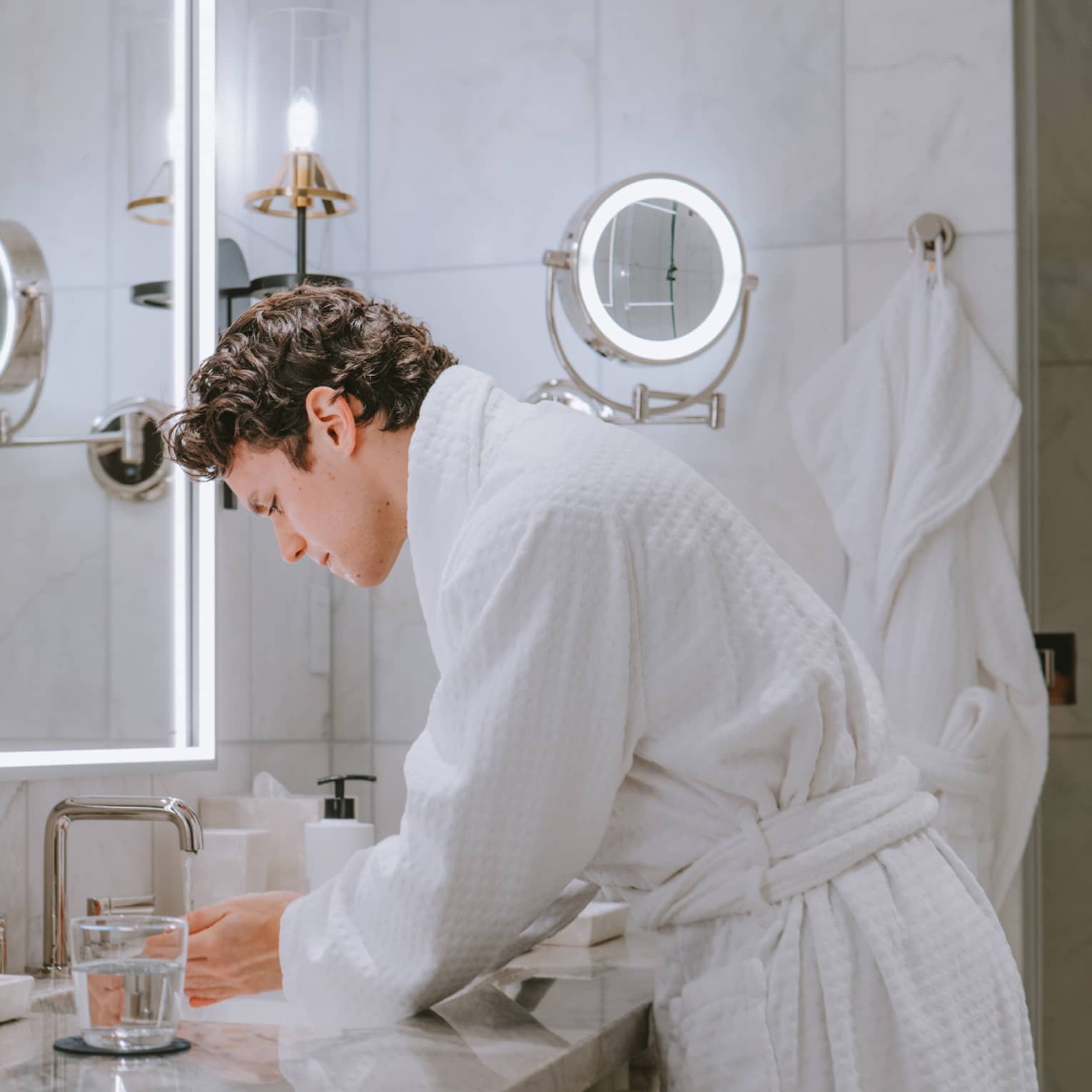 Person wearing a bath robe and washing their hands in the sink with a glass of water on the counter.