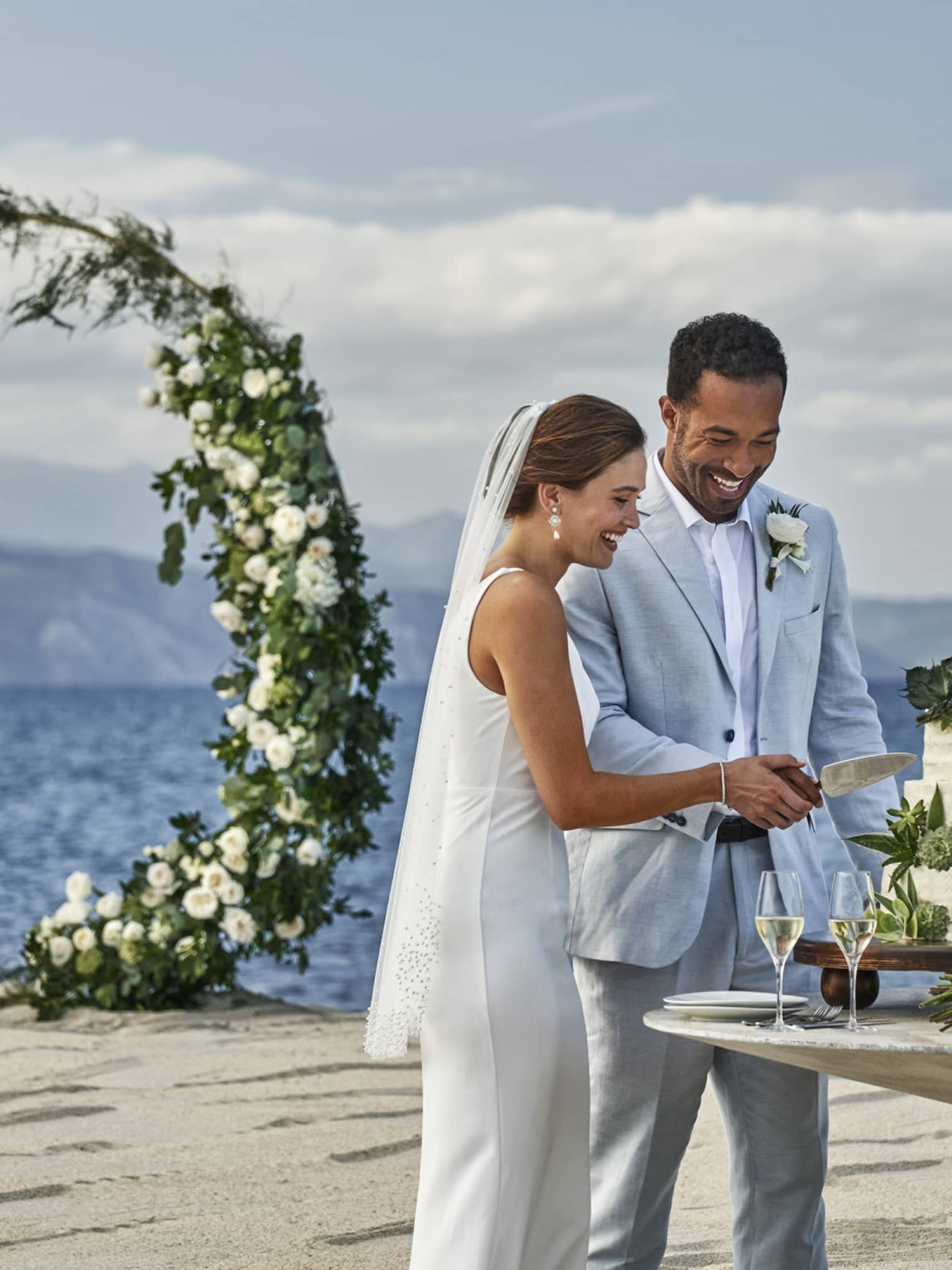 A bride and groom joyfully cut their wedding cake on a beautiful beach, surrounded by ocean waves.