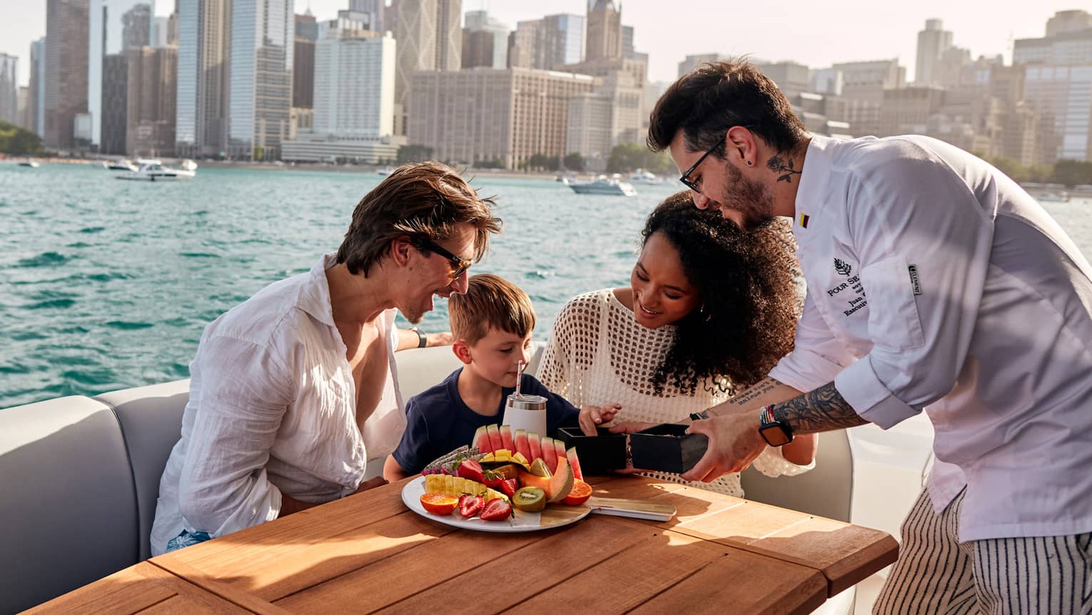 Parents watch their child sample from a dish offered by a chef as they sit at a table with sliced fruit on a boat deck.