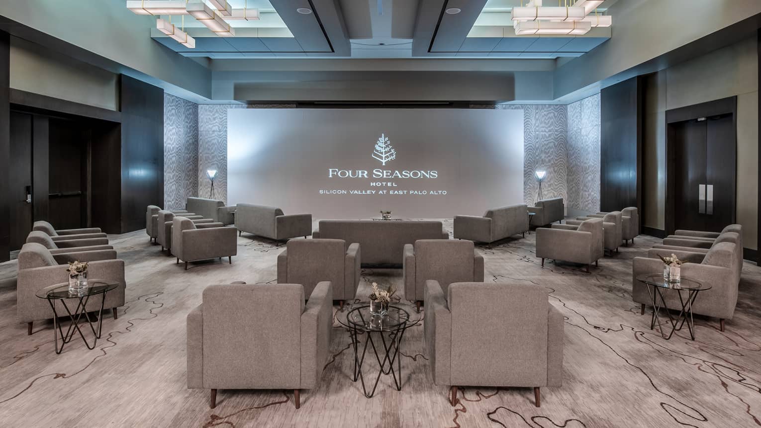 Gray plush chairs are angled towards a large projector in an indoor carpeted event space