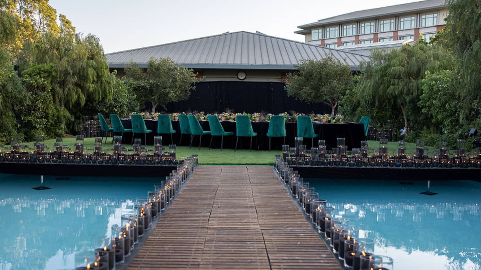 Wooden over-water walkway lined with candles in hurricane vases leads up to a platform where a large dining table set for a reception sits in the centre