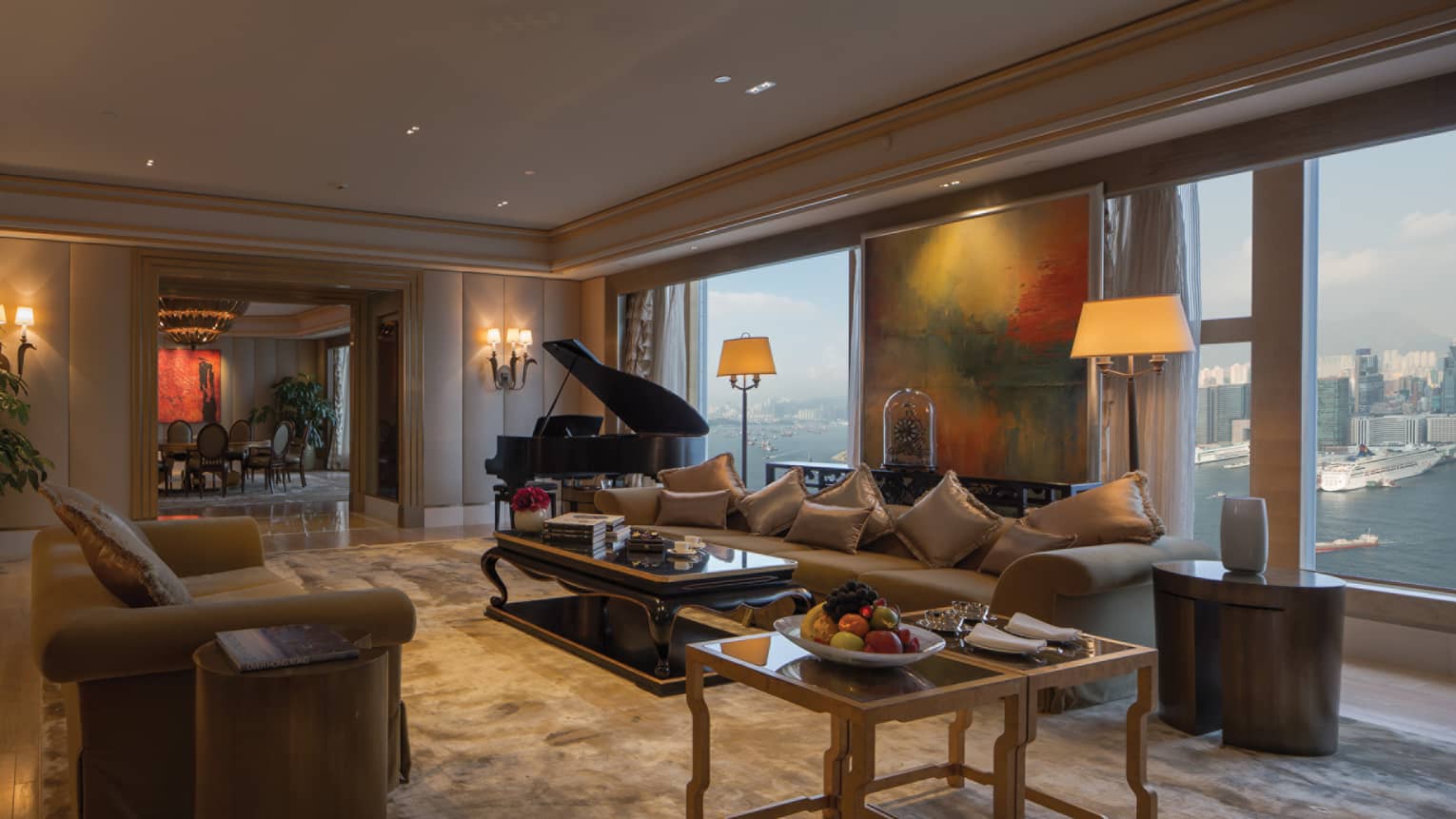 Luxurious living room with a grand piano, large windows with waterfront views, and a dining area in the background. A coffee table holds books. 