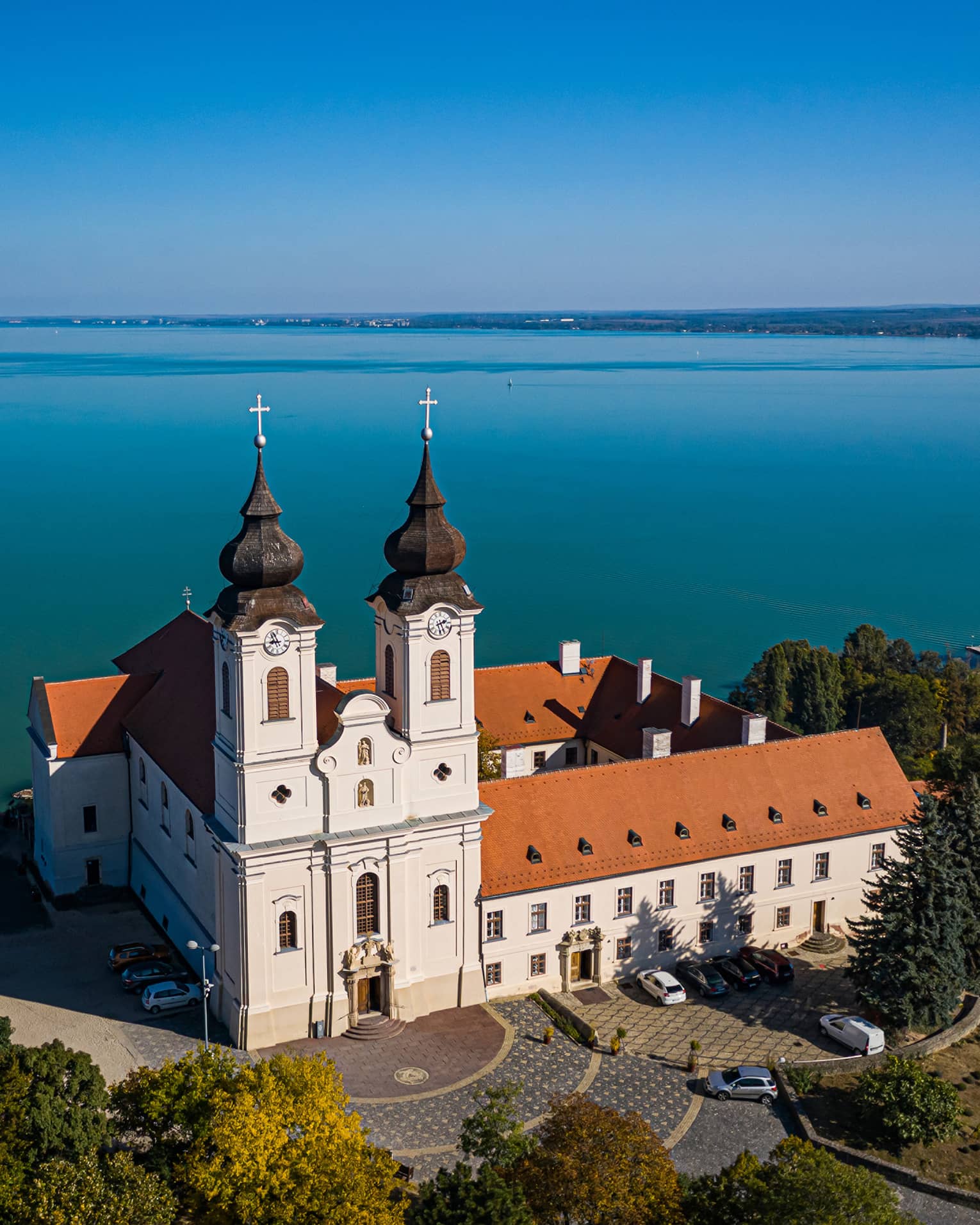 A Benedictine monastery, with white walls and a red tiled roof, sitting beside a large blue lake and surrounded by trees.