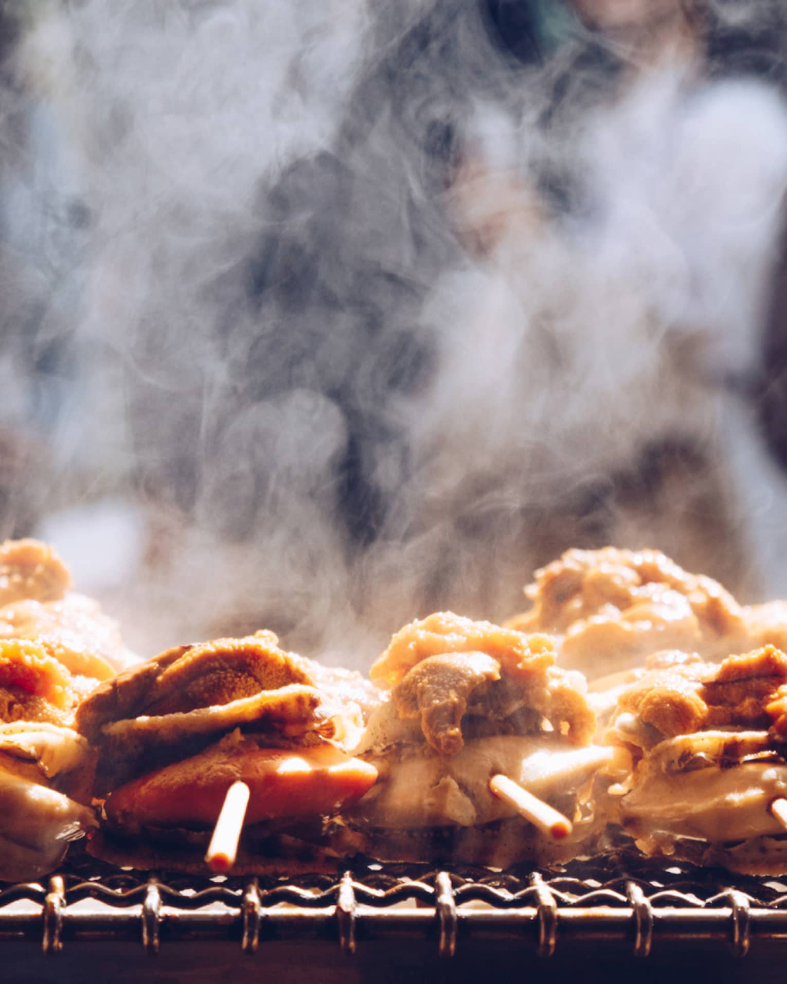 A close-up of steaming street food skewed on a stick, a line of patrons fill the background