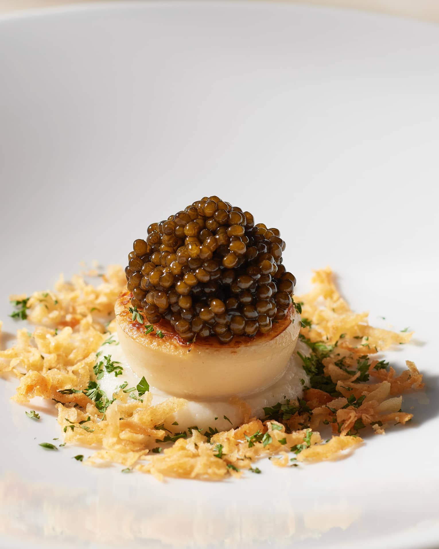 Round white plate with single scallop topped with caviar, surrounded by breadcrumbs and a smattering of green herbs