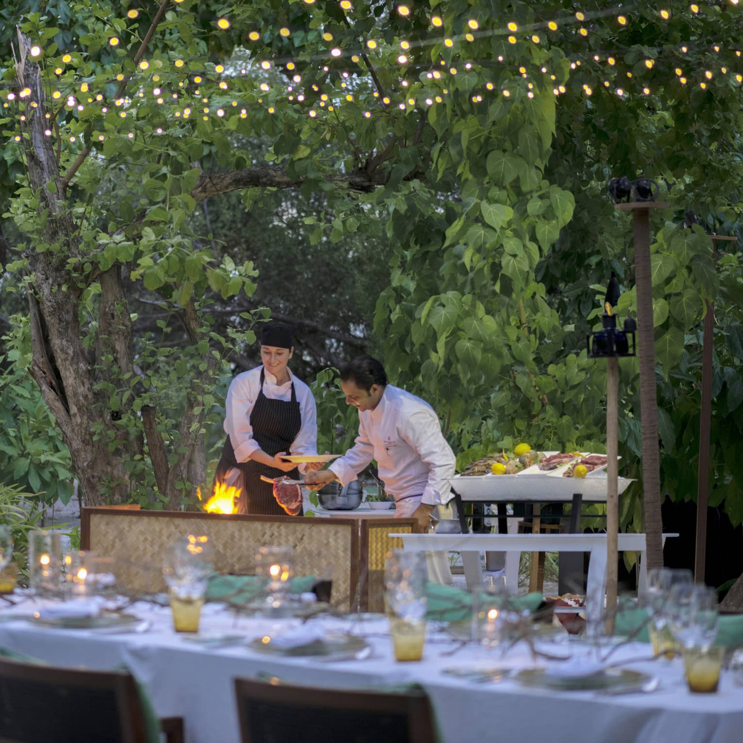 Chefs put large steak on flaming grill by long outdoor dining table, patio lights at night