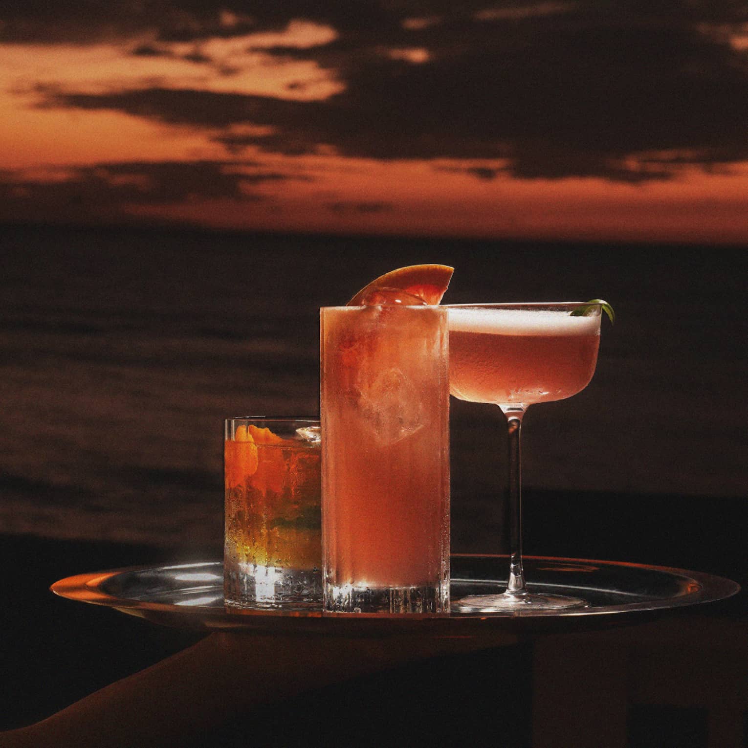 Server carries three cocktails on a tray with an orange-coloured sky in the background