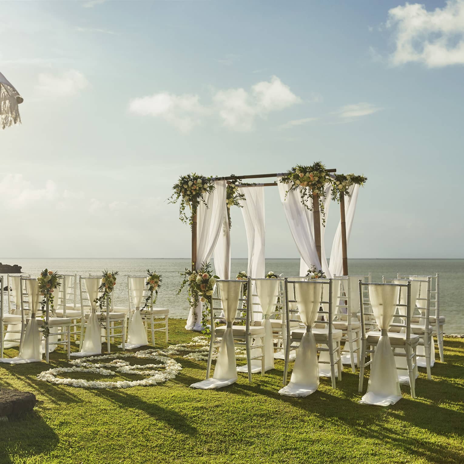Sun shines over wedding altar draped in white fabric, rows of chairs overlooking ocean