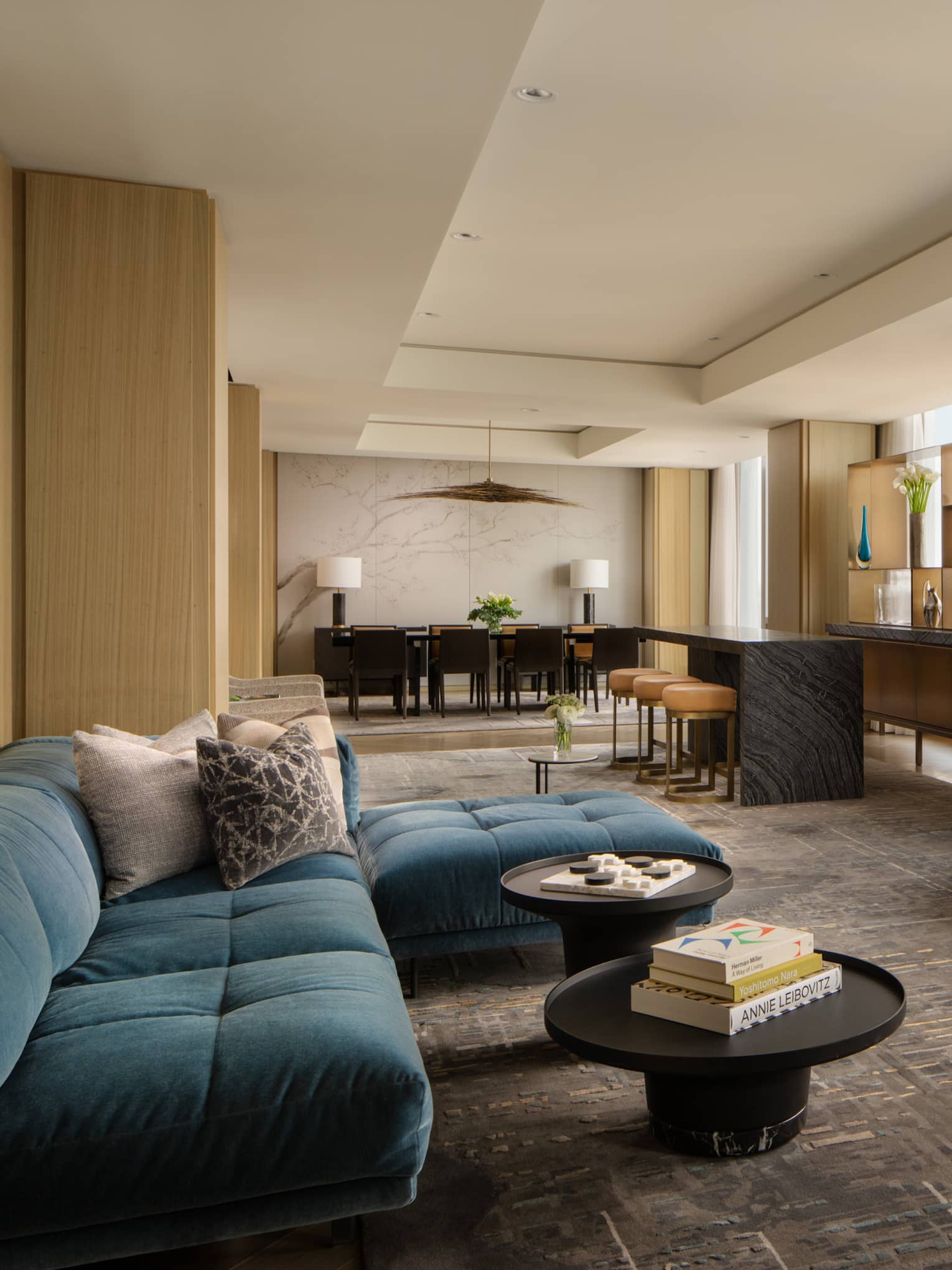 Luxury suite living area with blue sectional sofas, round coffee tables, bar with stools, dining table and floor-to-ceiling windows with city views