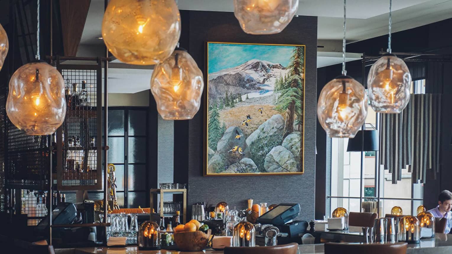 Amber-tinged glass lamps hang over bar in front of modern landscape painting in Goldfinch Tavern
