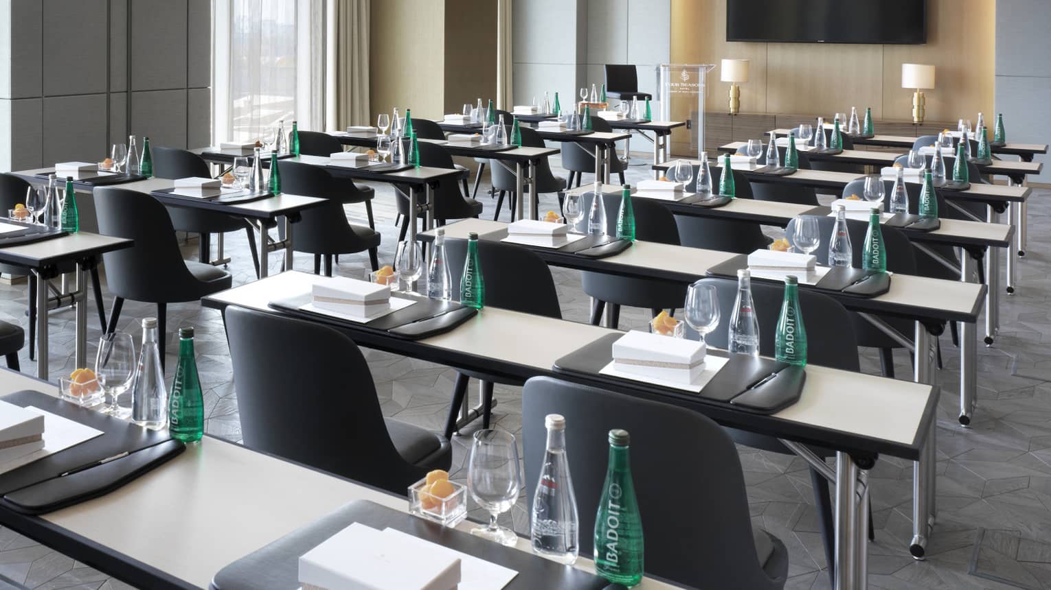 Two rows of desks with water, snacks, stationery and chairs facing TV in sleek boardroom
