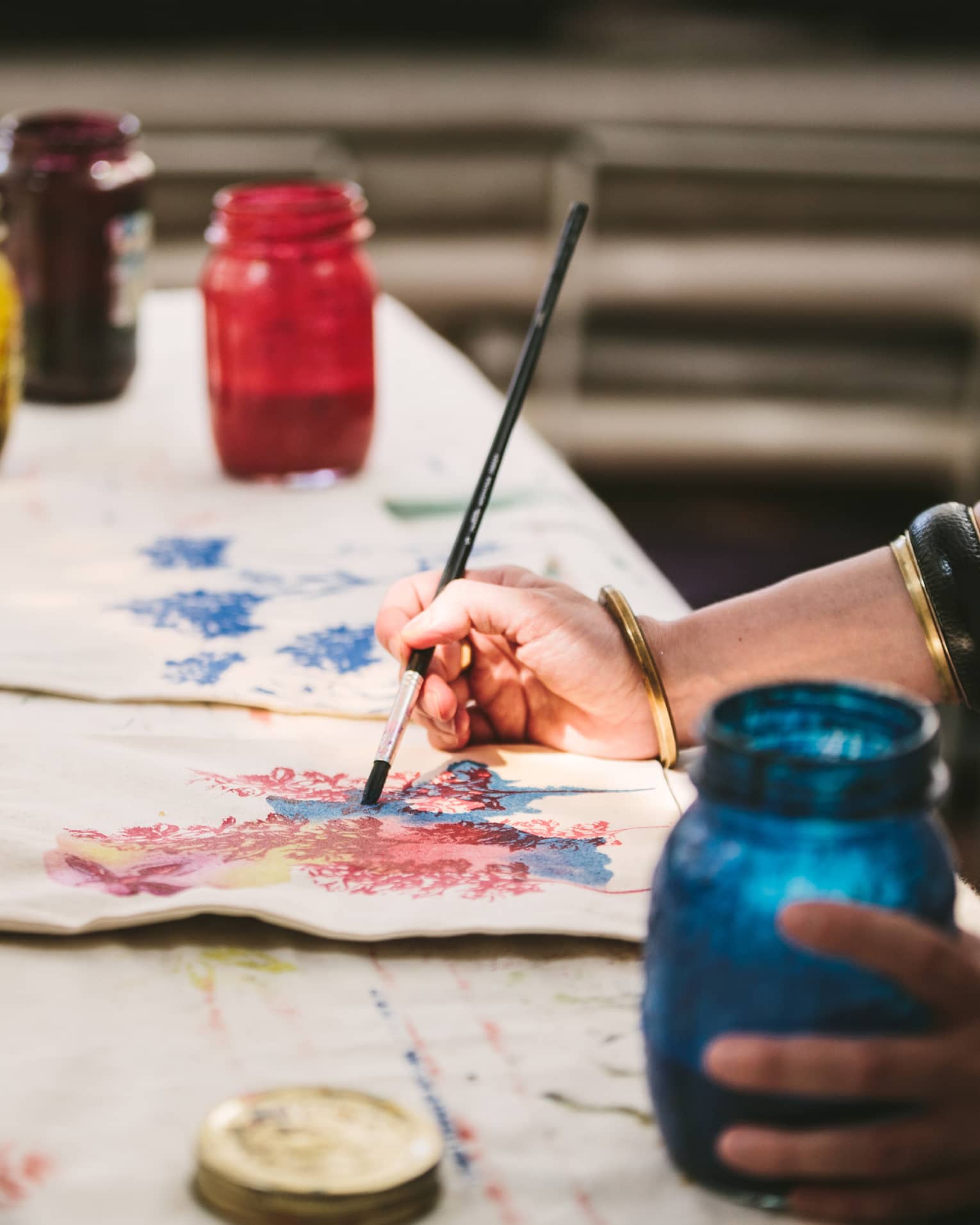An artist using mason jars filled with red, blue, yellow and black paint to paint images of flowers on canvas tote bags.