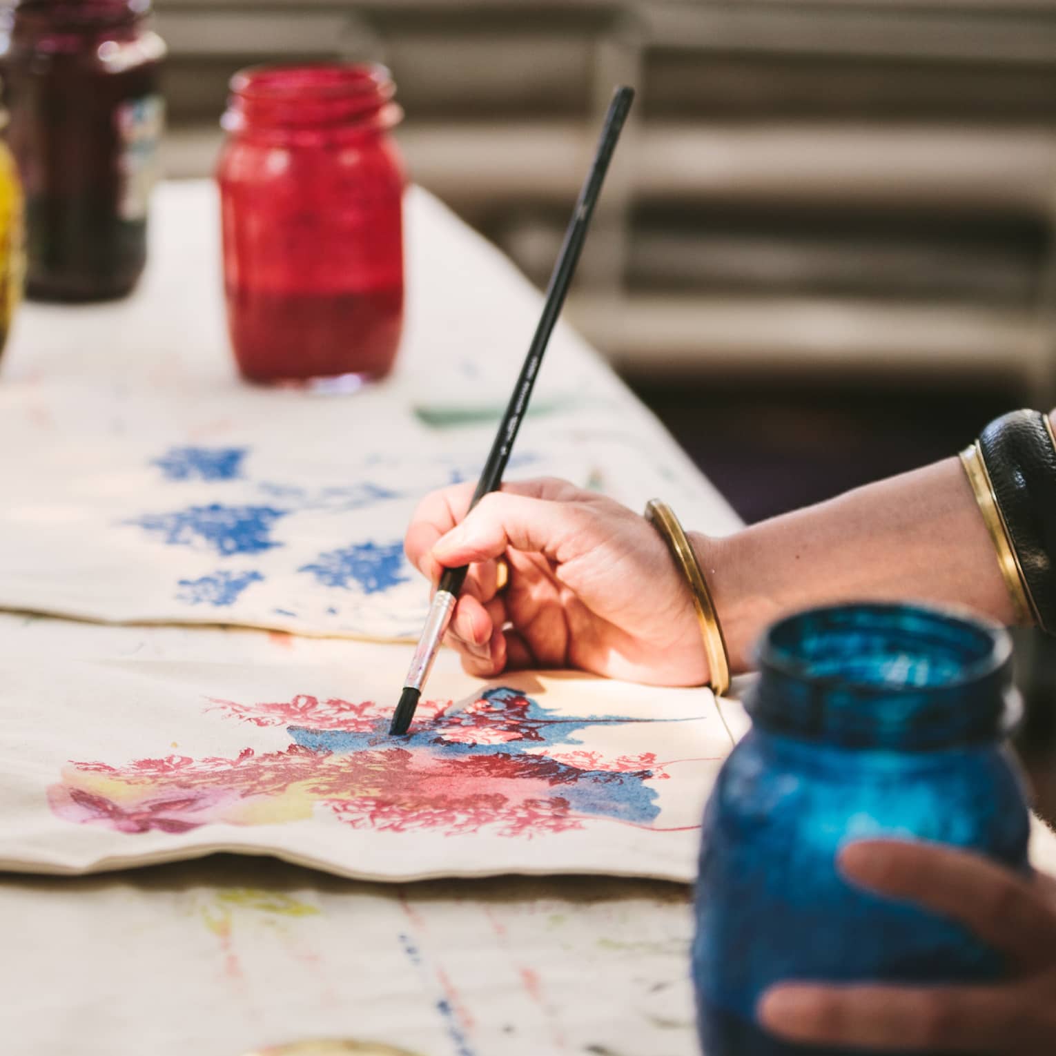 An artist using mason jars filled with red, blue, yellow and black paint to paint images of flowers on canvas tote bags.