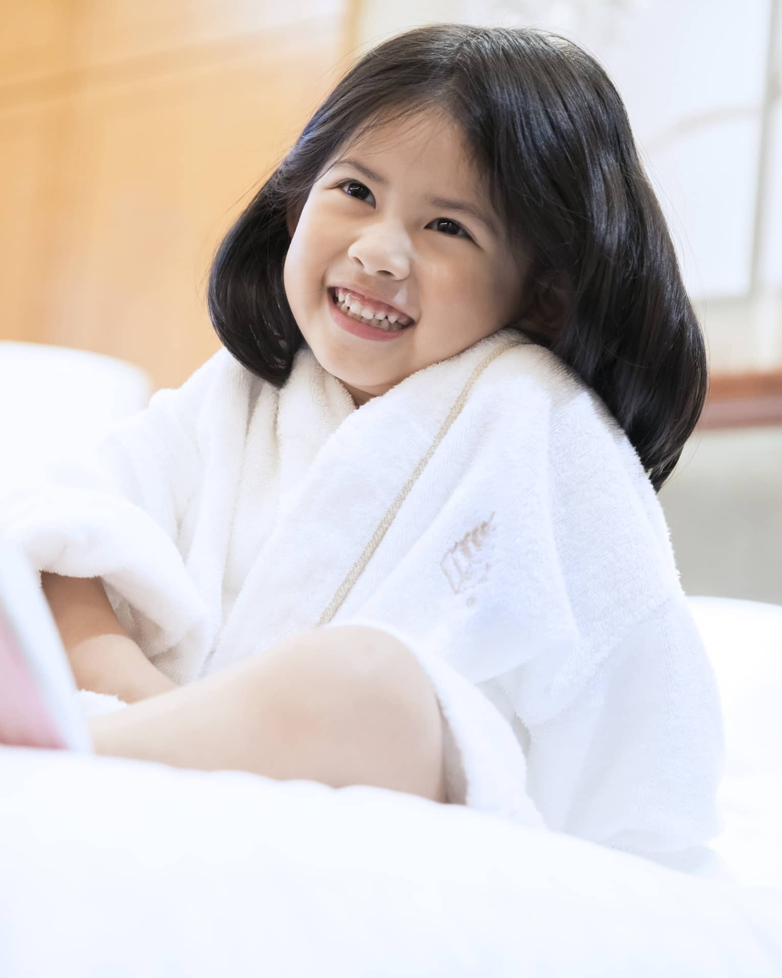 Smiling little girl wearing white bath robe and pink slippers sits on bed