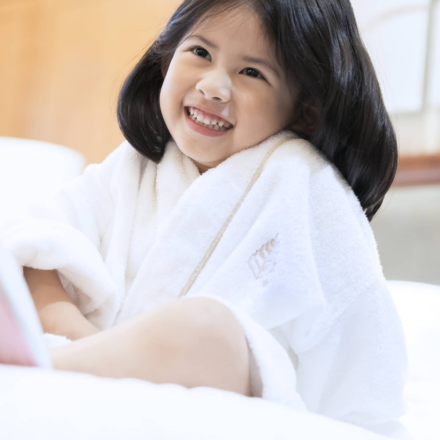 Smiling little girl wearing white bath robe and pink slippers sits on bed