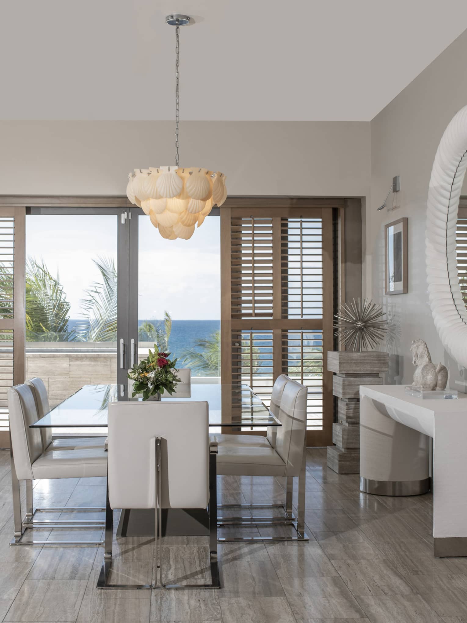 Luxury resort penthouse dining area with a glass table, white leather chairs, coastal decor and large windows with wooden shutters offering an ocean view