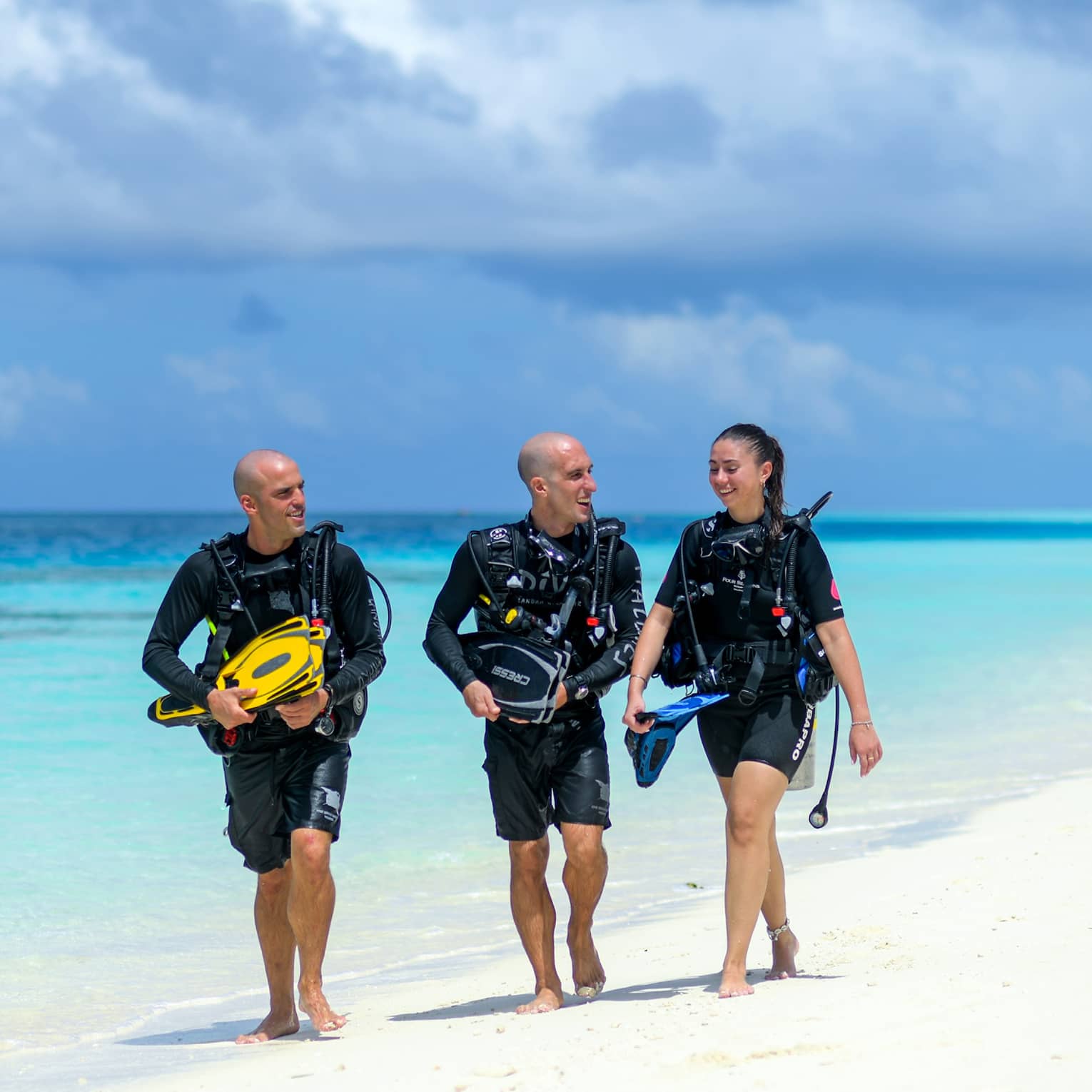 Three smiling people wearing scuba gear, carrying their flippers, walk on a powdery-sand beach at a pale blue ocean's edge.