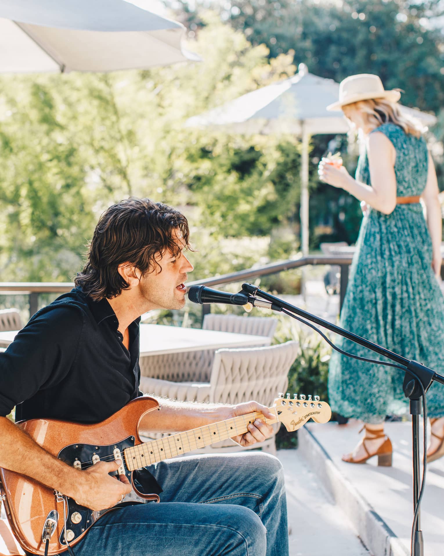 A man sitting and playing guitar while a woman in a green dress walks behind him.