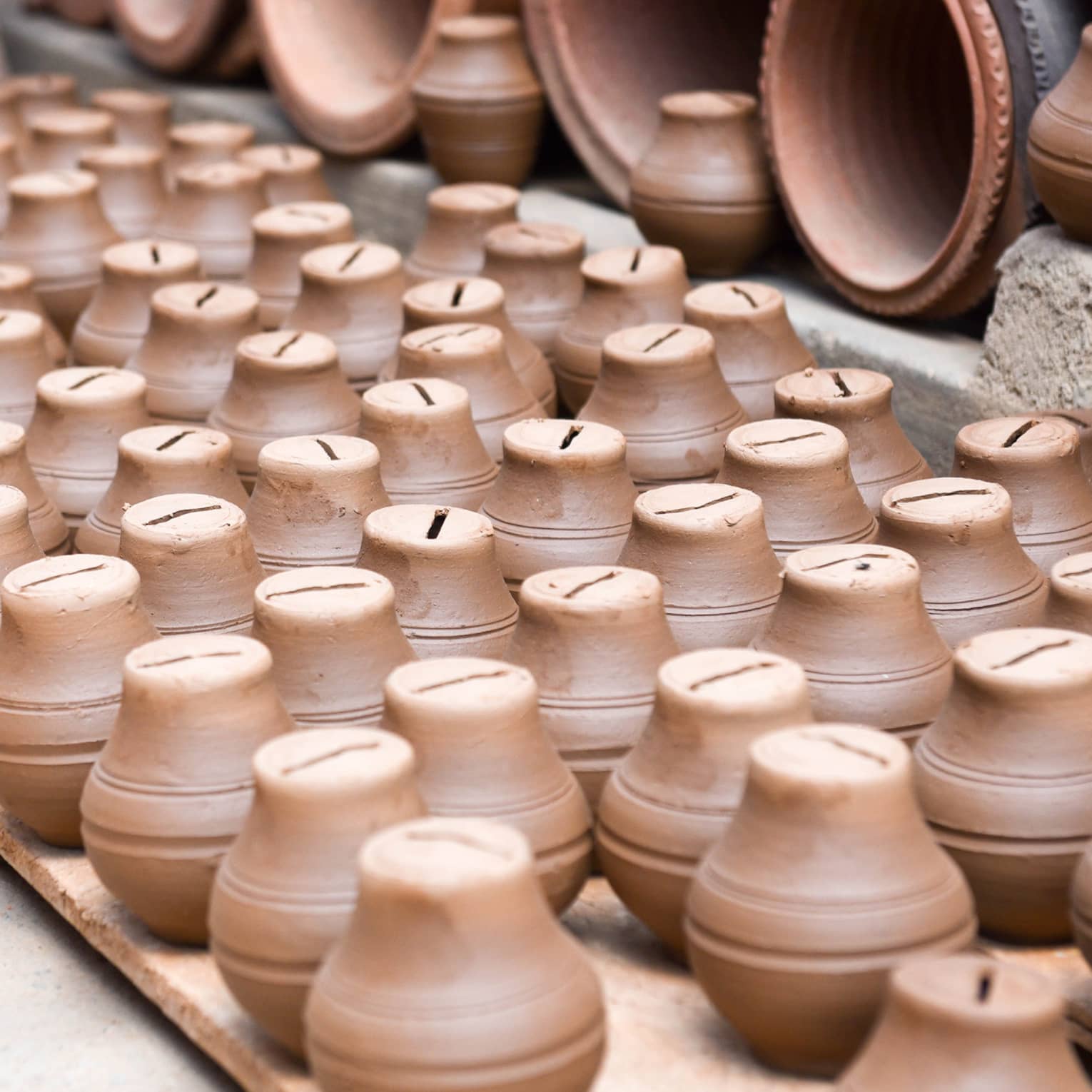 A collection of small, brown clay piggy banks neatly arranged on wooden pallets, surrounded by larger pottery items in the background, creating a display of handcrafted ceramics.