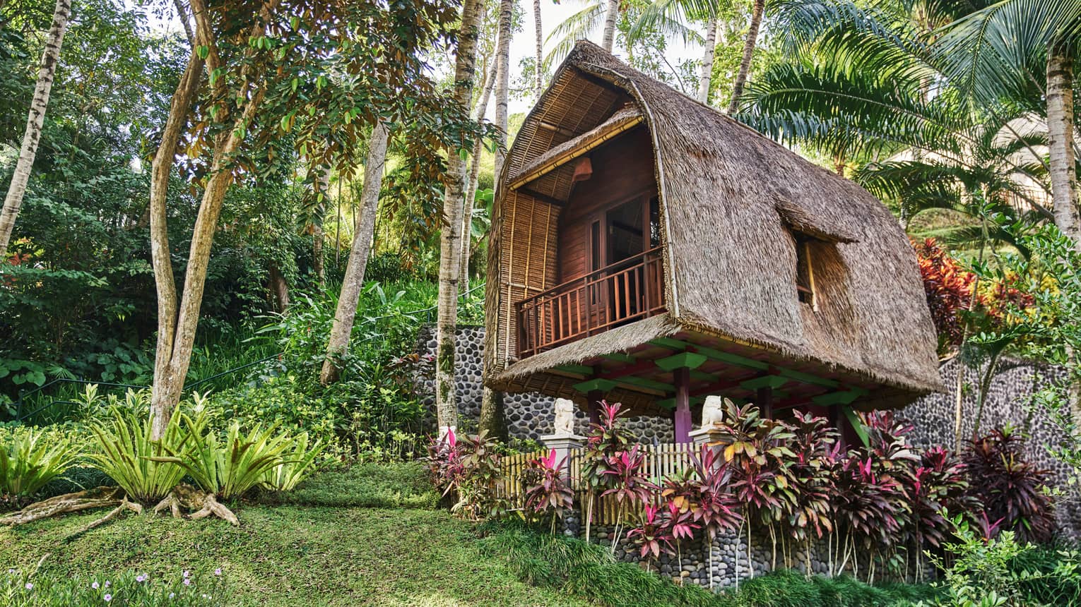 Exterior view of rice barn tree house with curved, A-frame structure, thatched roof, surrounded by tropical foliage