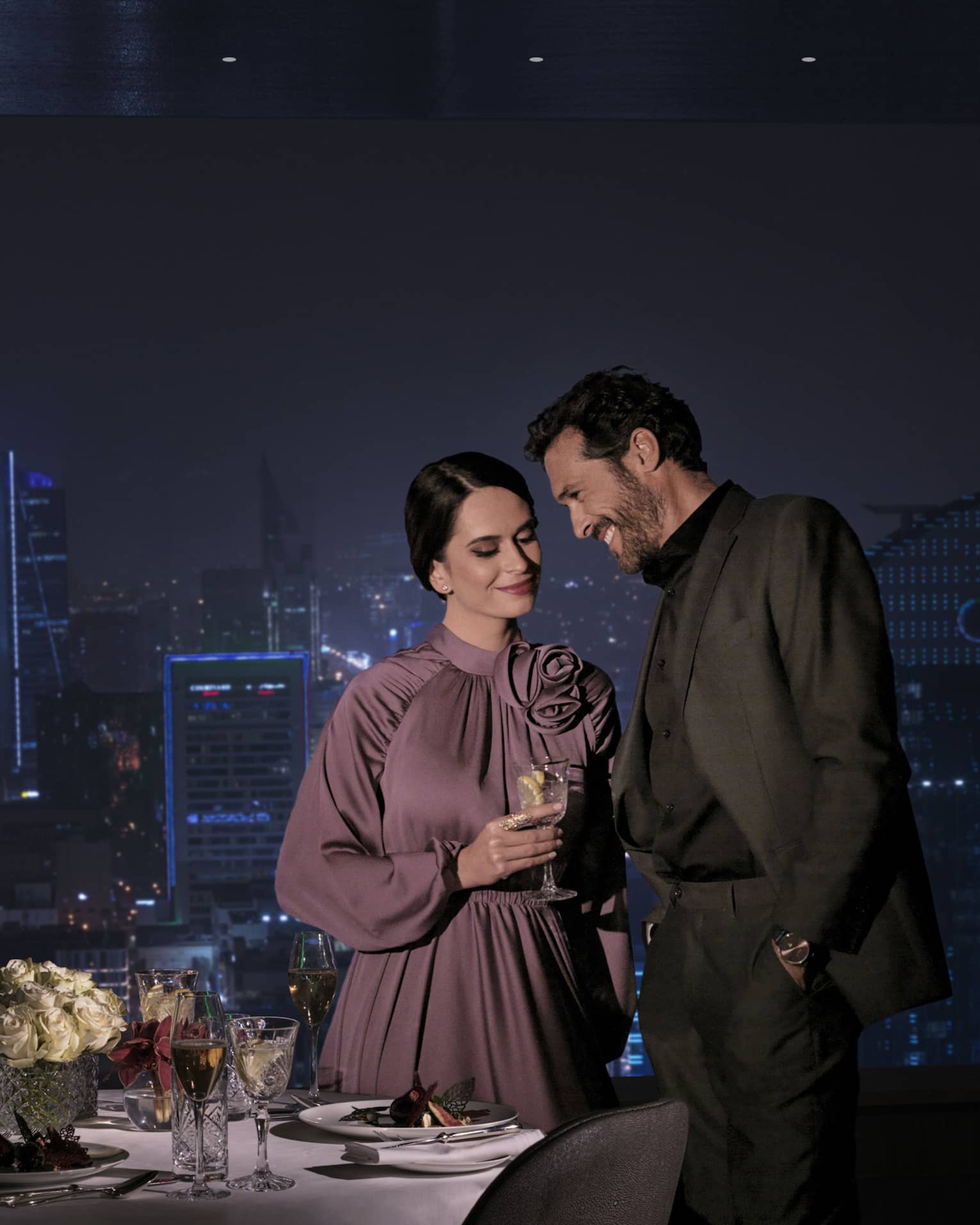 A beautiful view of the cityscape at night, a loving couple stands in front of the view with a romantically set dinner table.