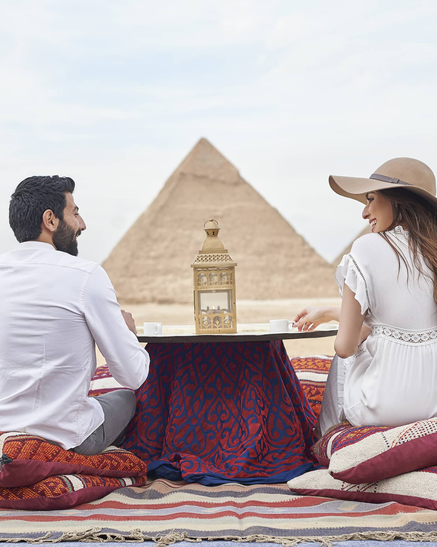 Back of couple sitting on pillows by small dining table in desert looking at pyramids