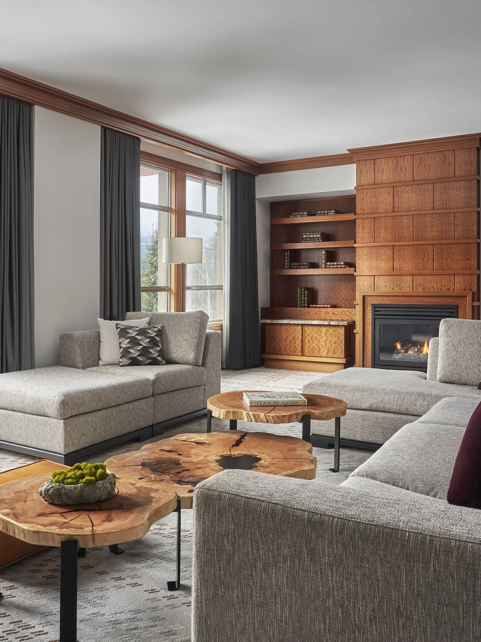 Spacious and stylish Blackcomb Suite at Four Seasons Whistler featuring a sophisticated design with large windows offering a view of the mountains. The suite includes two distinct seating areas: one with a large gray sectional sofa adorned with maroon pillows, and another with a beige daybed by the window. Wooden coffee tables, elegant carpeting, and a built-in wooden shelving unit with a fireplace and a flat-screen TV add to the luxurious feel of the room.,Spacious and stylish Blackcomb Suite at Four Seasons Whistler featuring a sophisticated design with large windows offering a view of the mountains. The suite includes two distinct seating areas: one with a large gray sectional sofa adorned with maroon pillows, and another with a beige daybed by the window. Wooden coffee tables, elegant carpeting, and a built-in wooden shelving unit with a fireplace and a flat-screen TV add to the luxurious feel of the room.