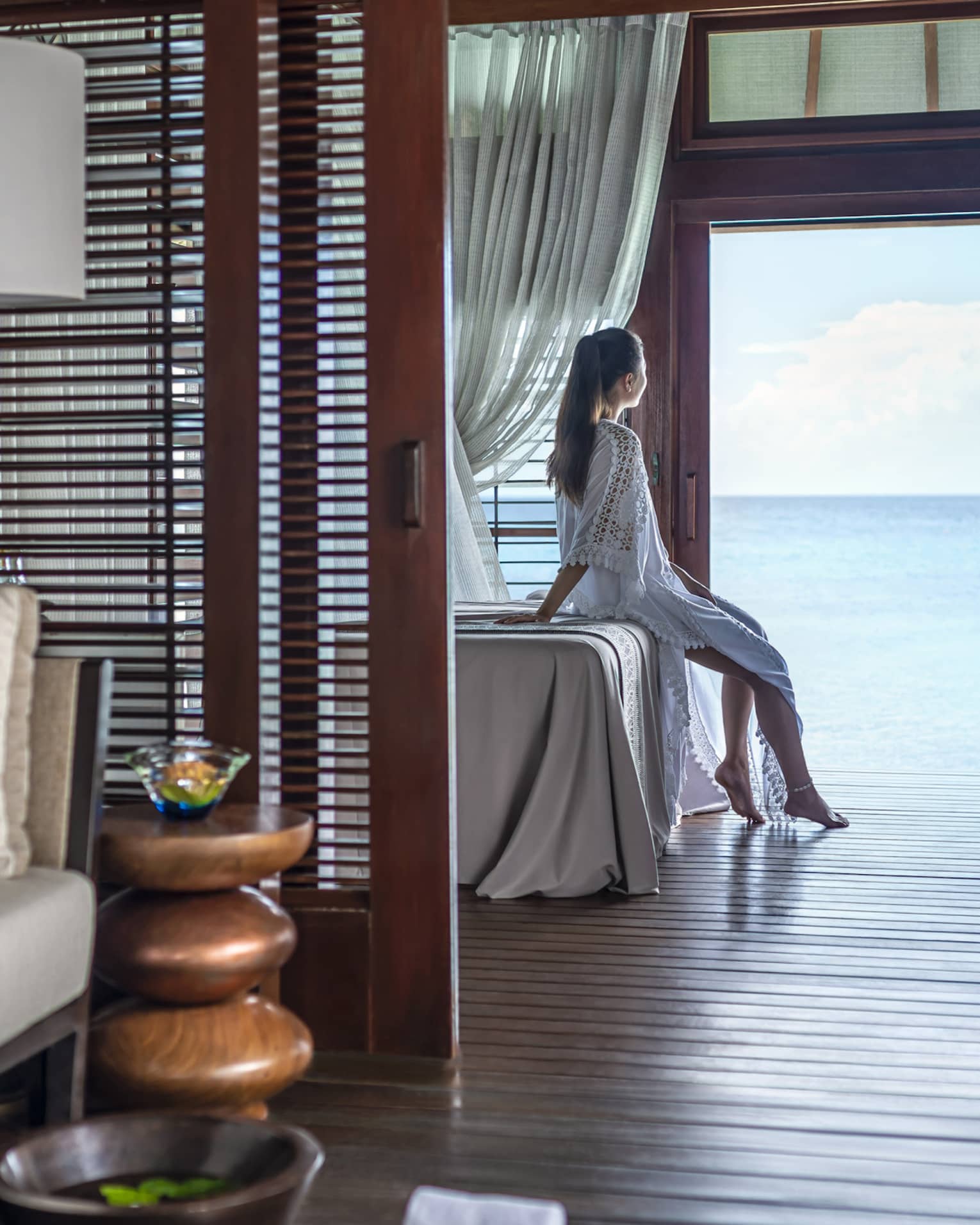 A woman leans on a massage bed on the far side of a spa treatment room and looks out through a large doorway onto the vast blue ocean