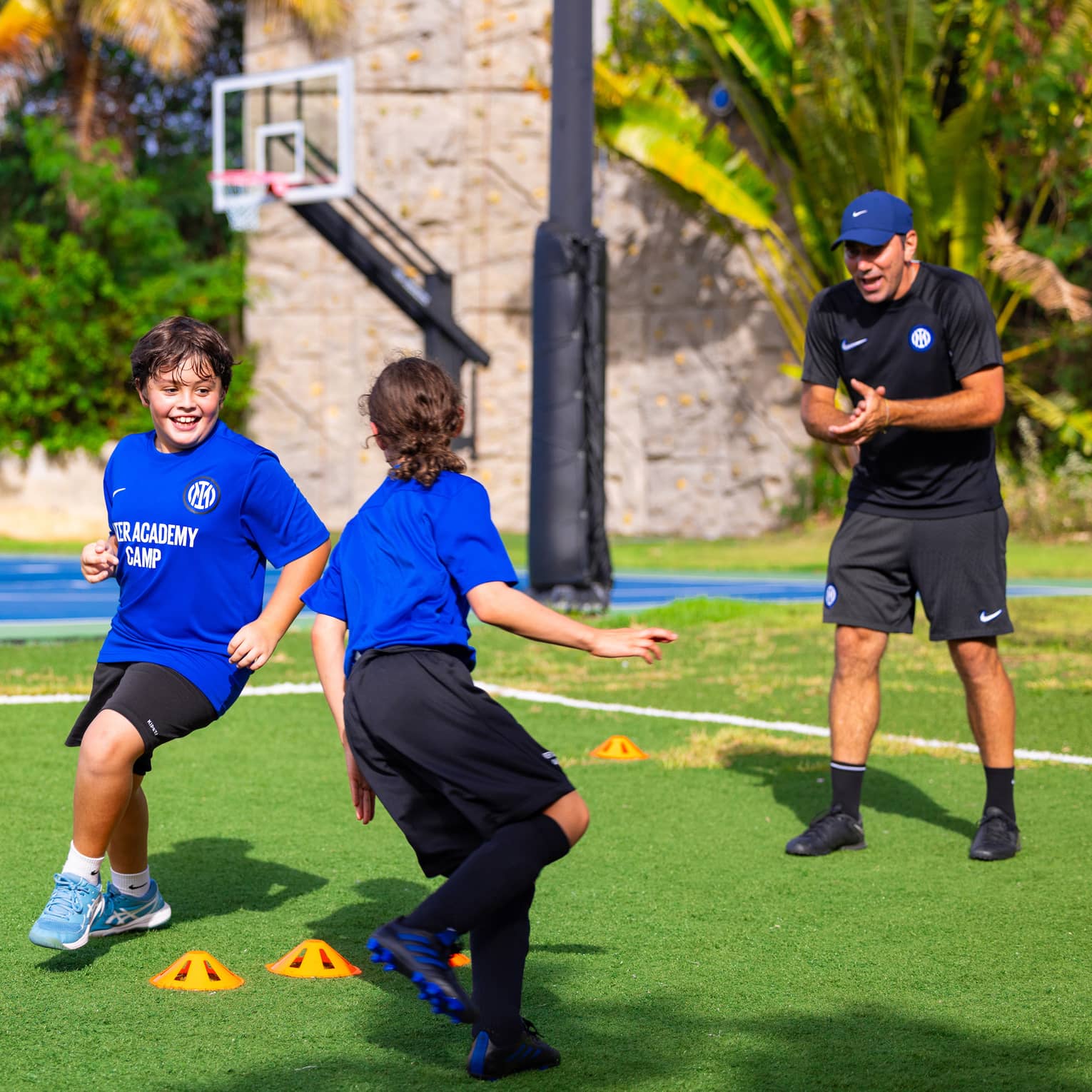 Soccer coach talking to two young guests exercising outside with a basketball court in the background.