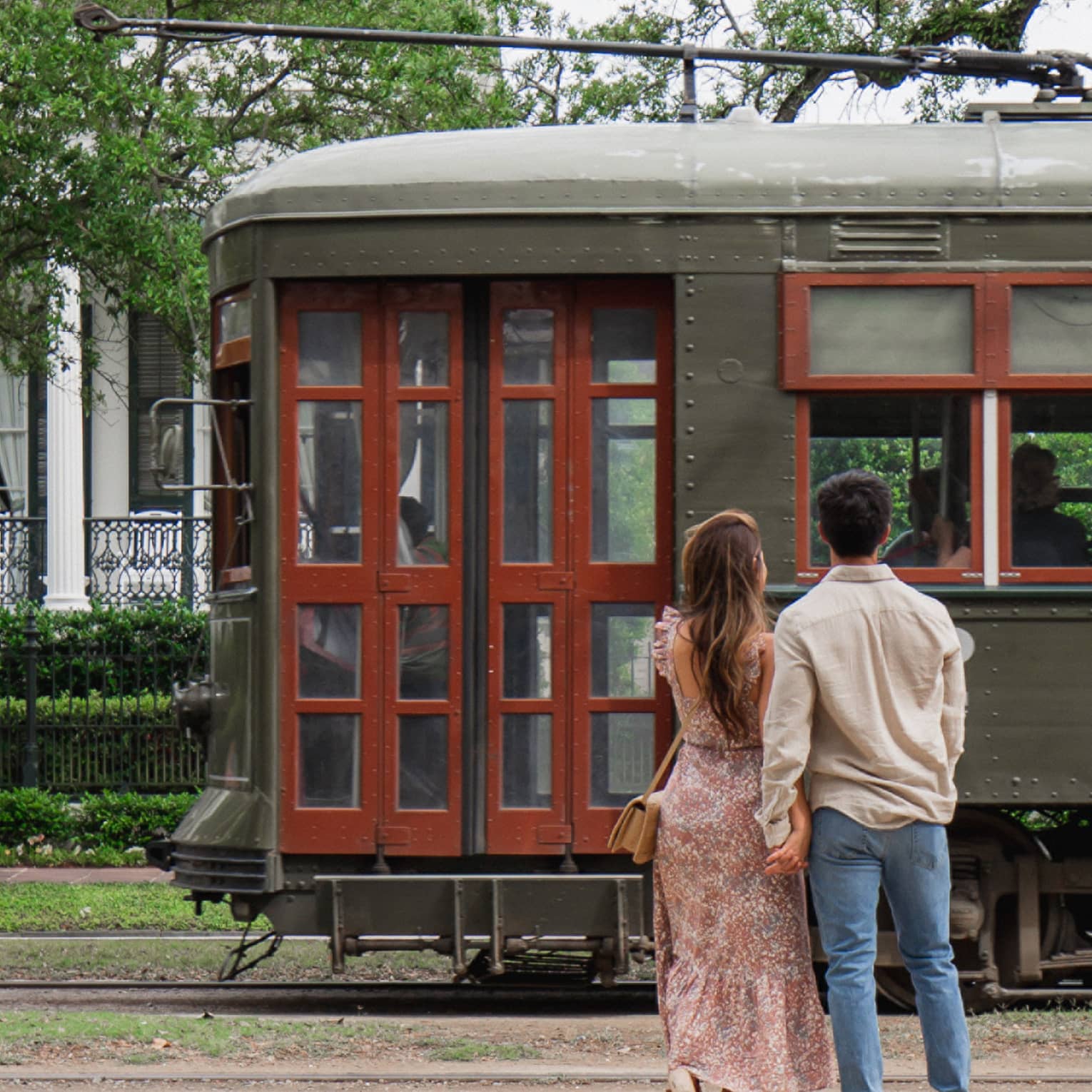 Woman and man hold hands in front of old-fashioned streetcar on New Orleans street, white house in background