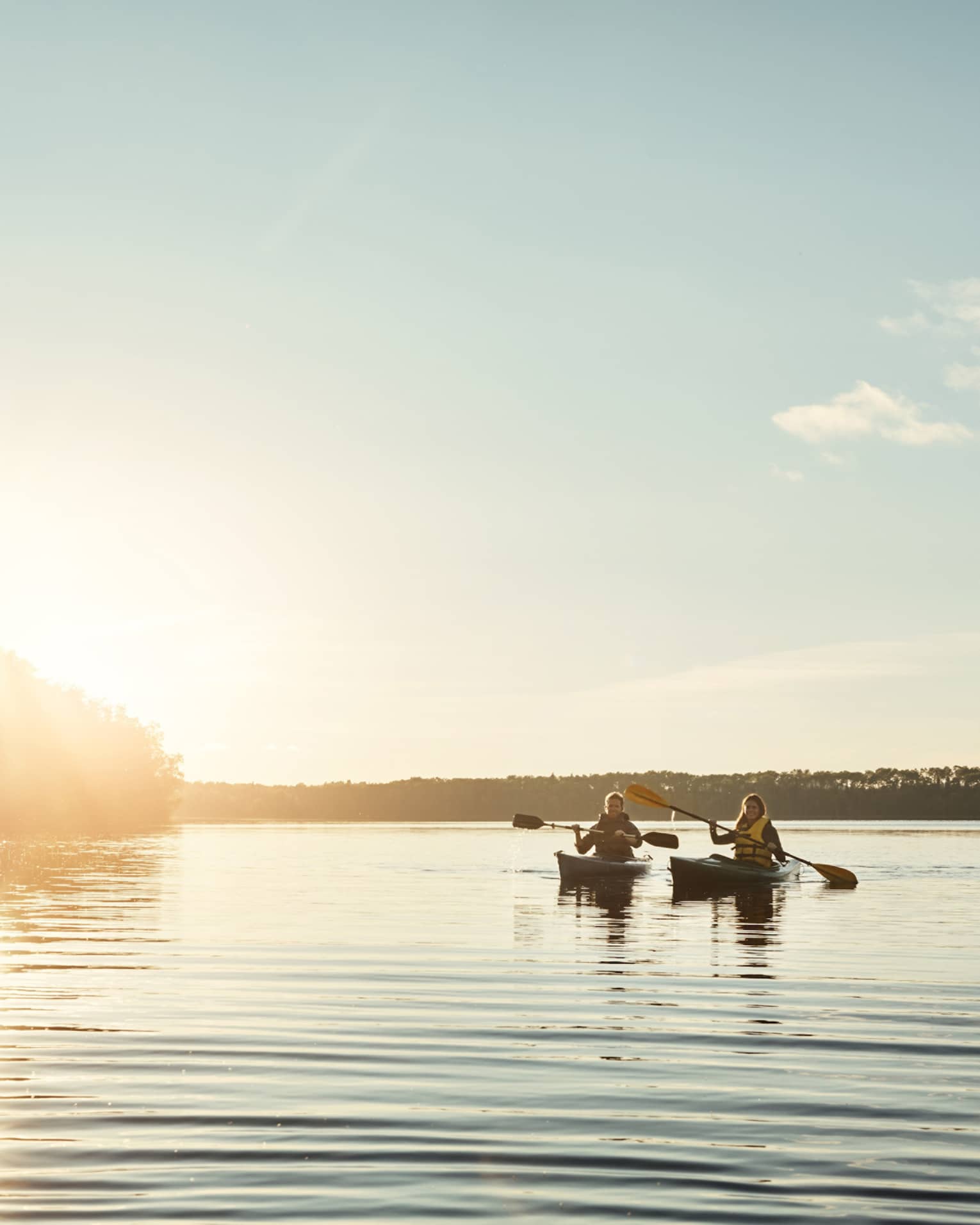Two people kayaking through a body of water flanked by trees, both silhouetted by the setting sun