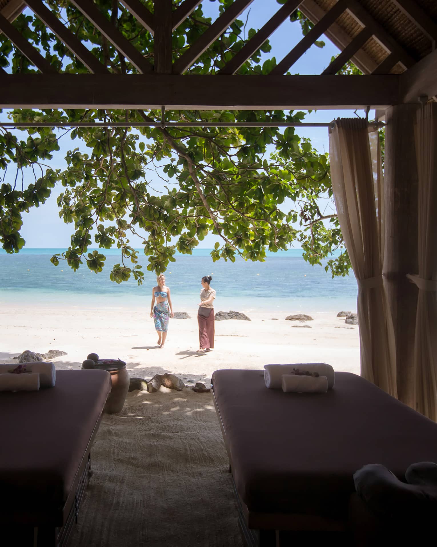 Couples massage beds under cabana in front of two women walking on white sand beach