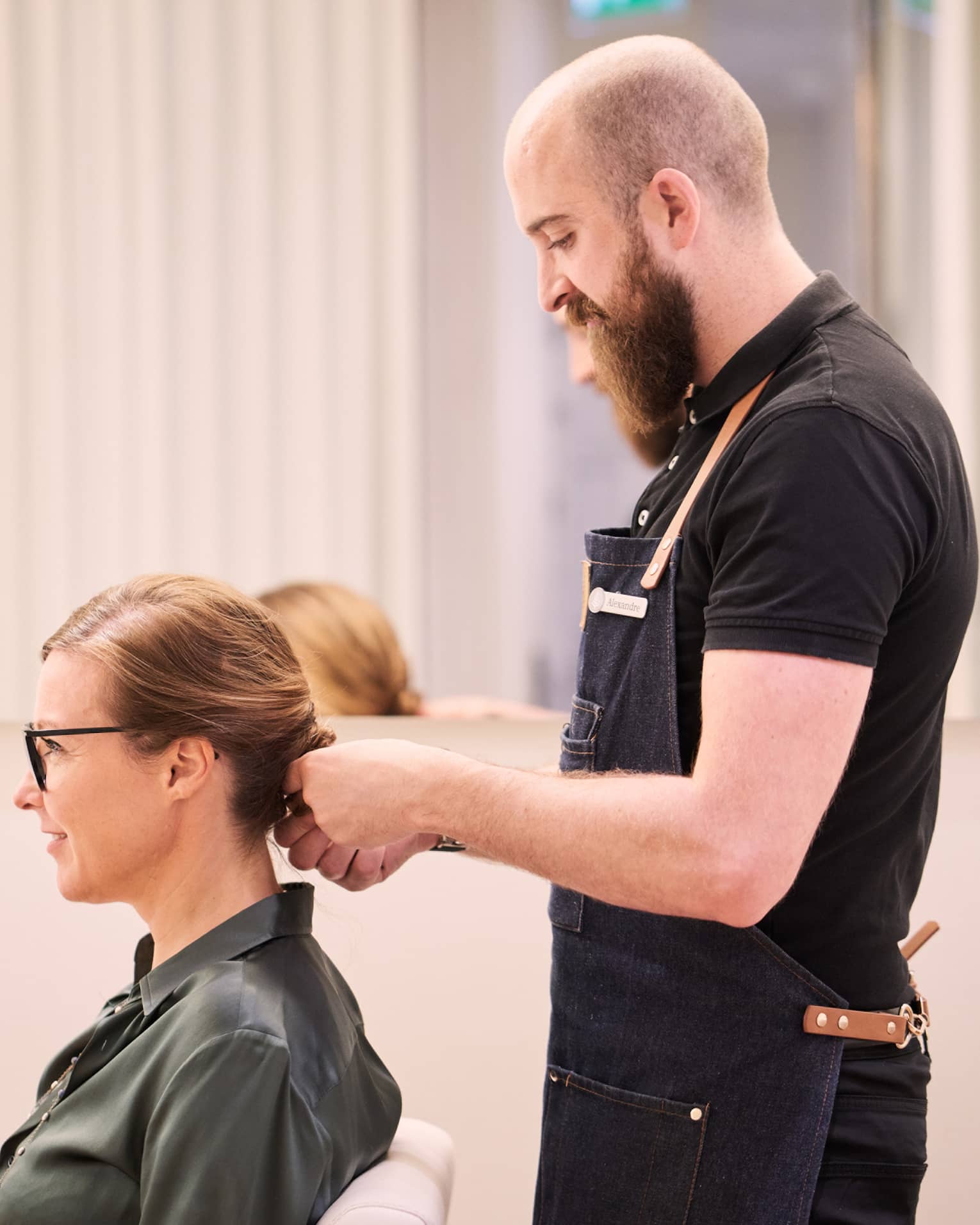 Woman sits in salon chair as hair stylist with long beard holds her hair
