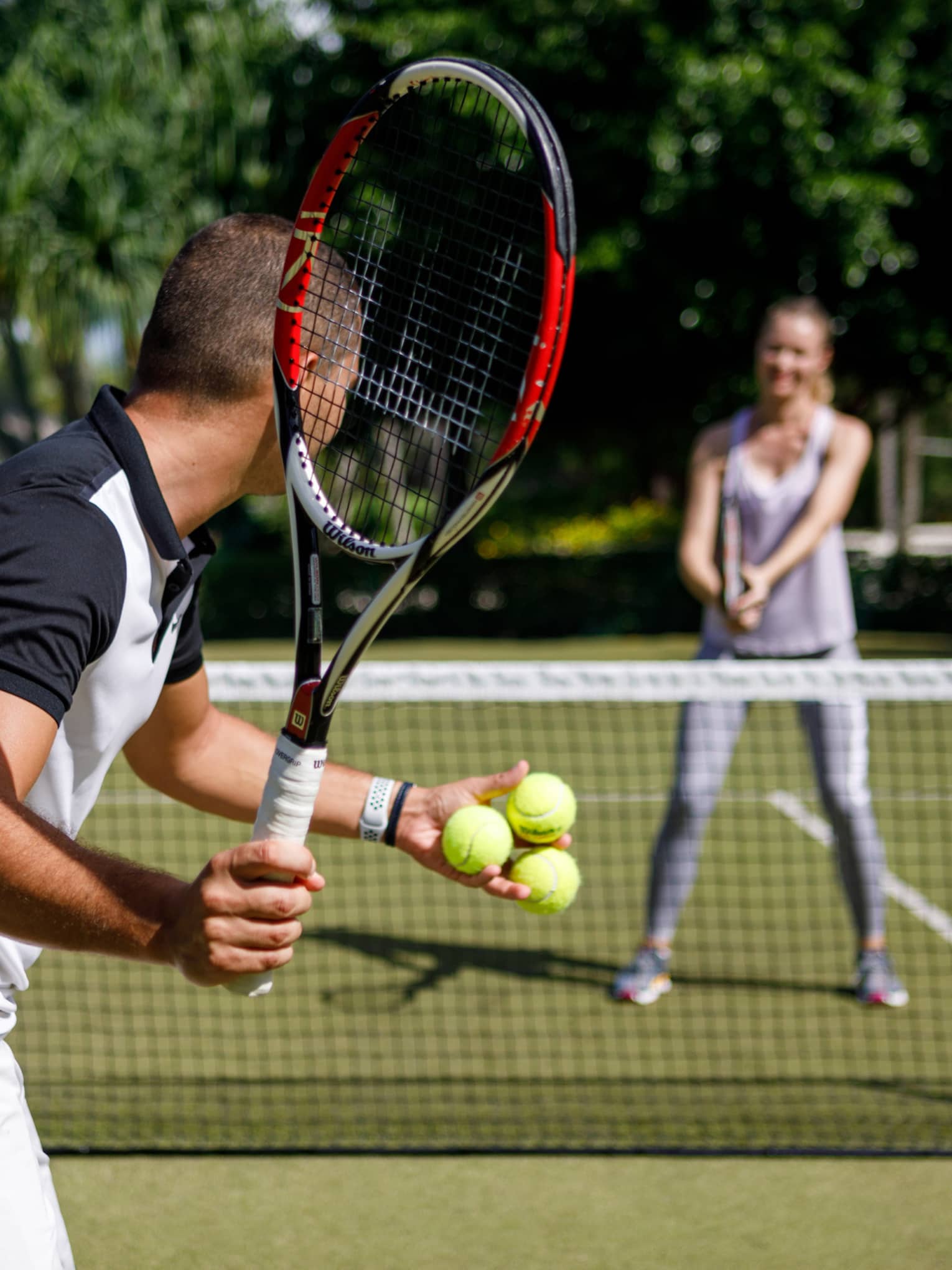 Couple playing tennis on court, man prepares to serve tennis balls to woman ready to swing