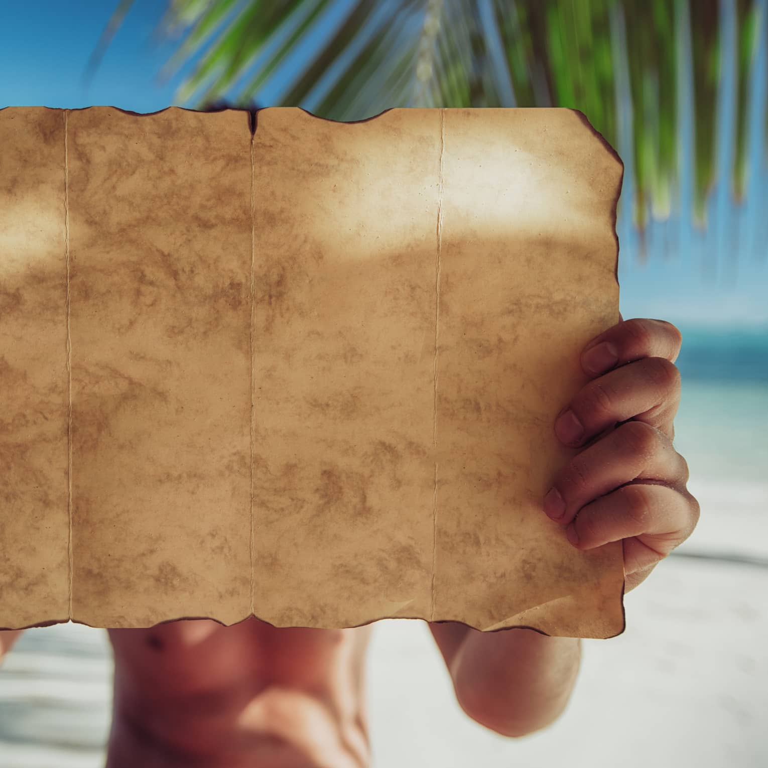 A close-up of a piece of parchment paper, yellow with age and burnt around the edges, being examined on a sandy beach.