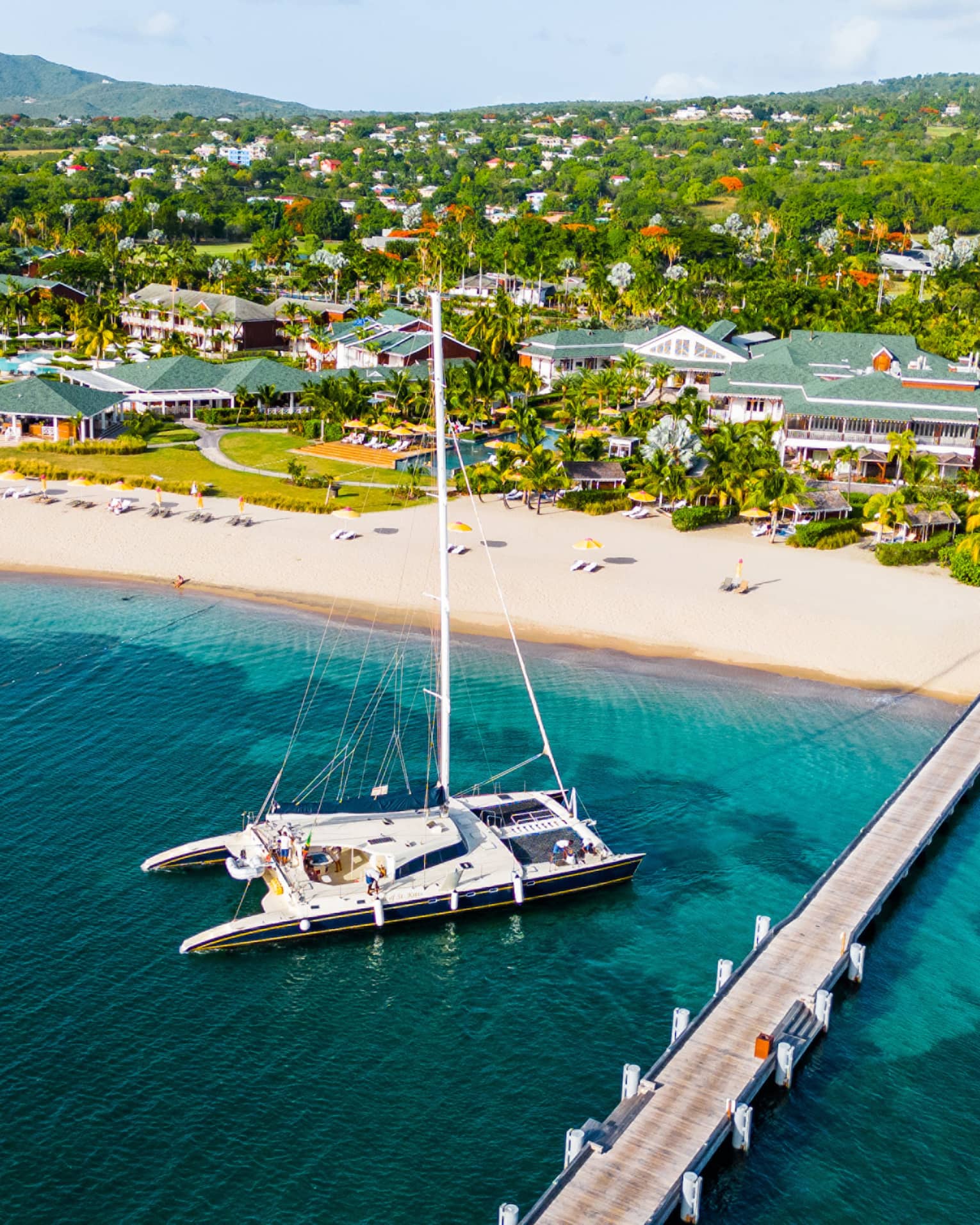 Aerial view of catamaran anchored near a dock reaching out from a beach with a sprawling resort along the coastline set among the trees