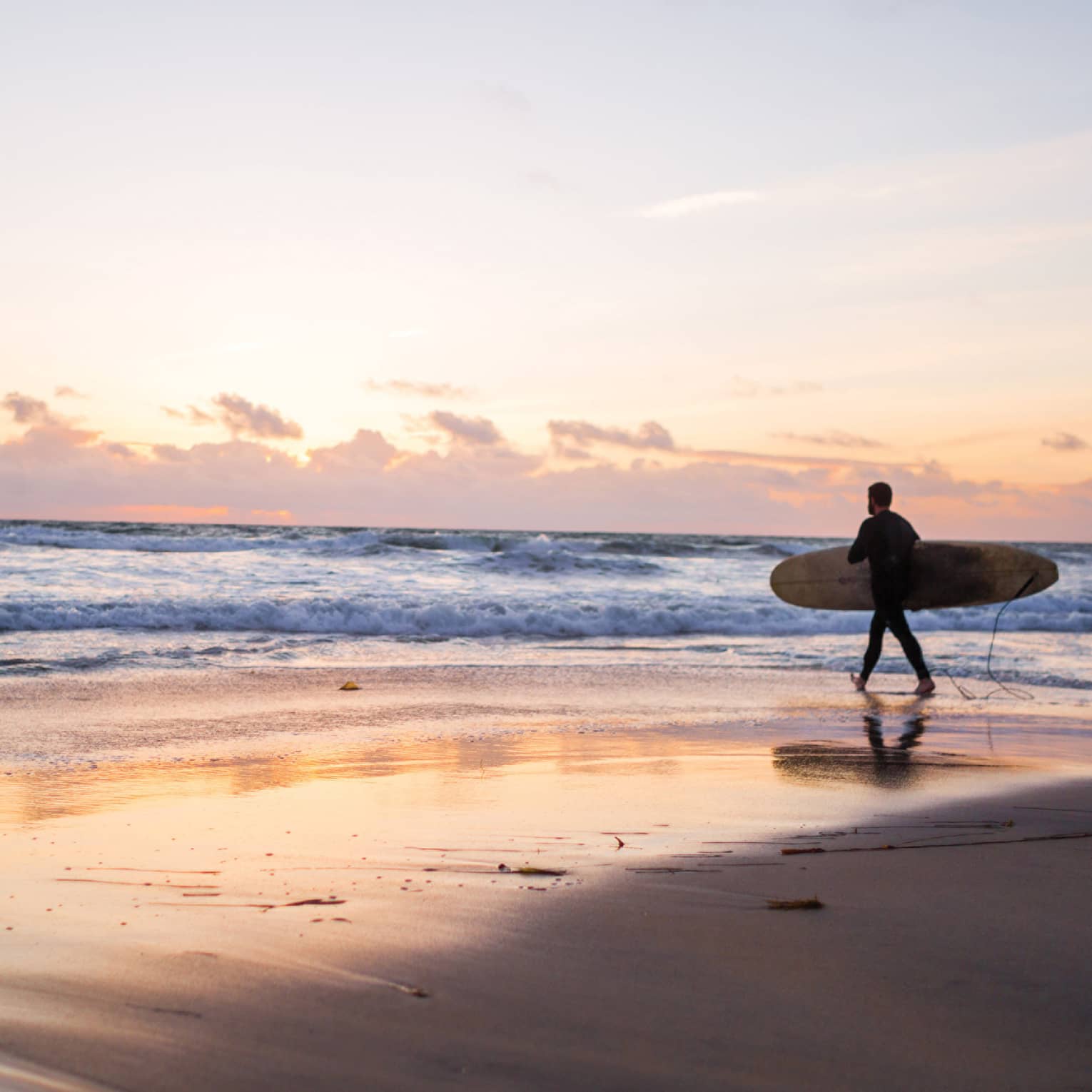 Under an expansive sky at sunset, clouds receding on the horizon, a surfer with a board heads into the glimmering ocean.