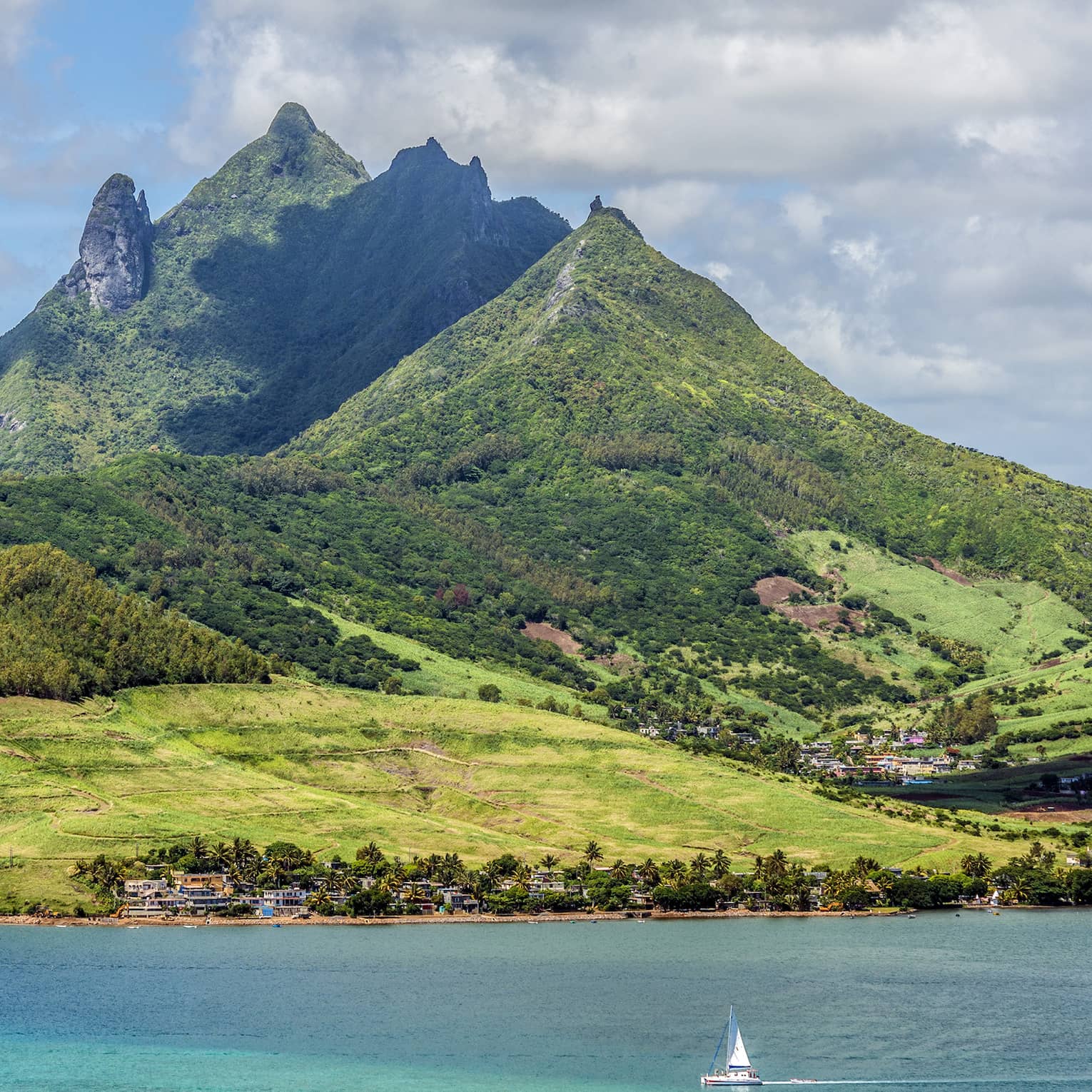 Island of lush, forested, multi-hued jagged hills over turquoise waters, a sailboat in the foreground dwarfed by the hills.