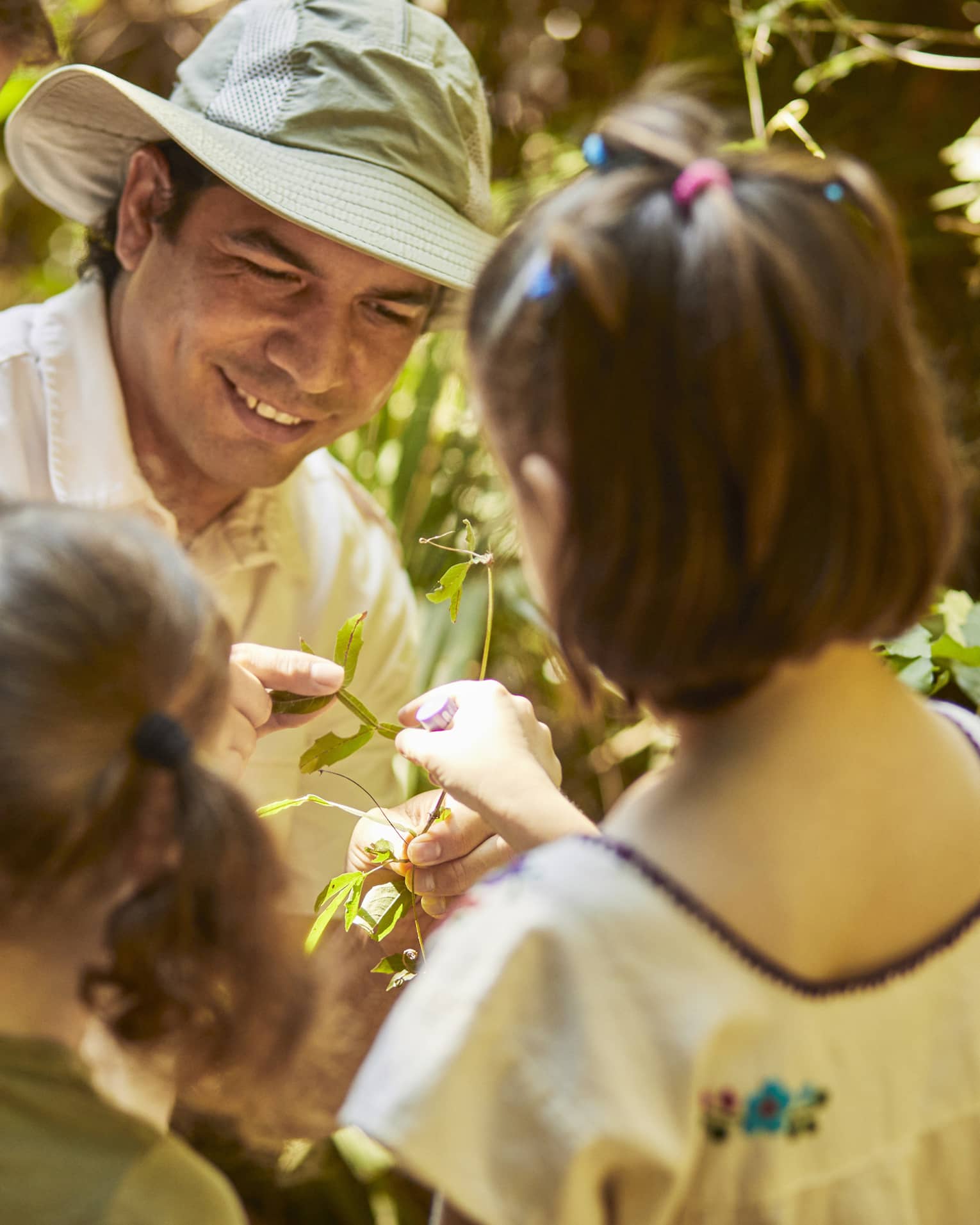 Three children gather around a guide wearing a sun hat as he shows them a plant. One child reaches to touch the leaves.