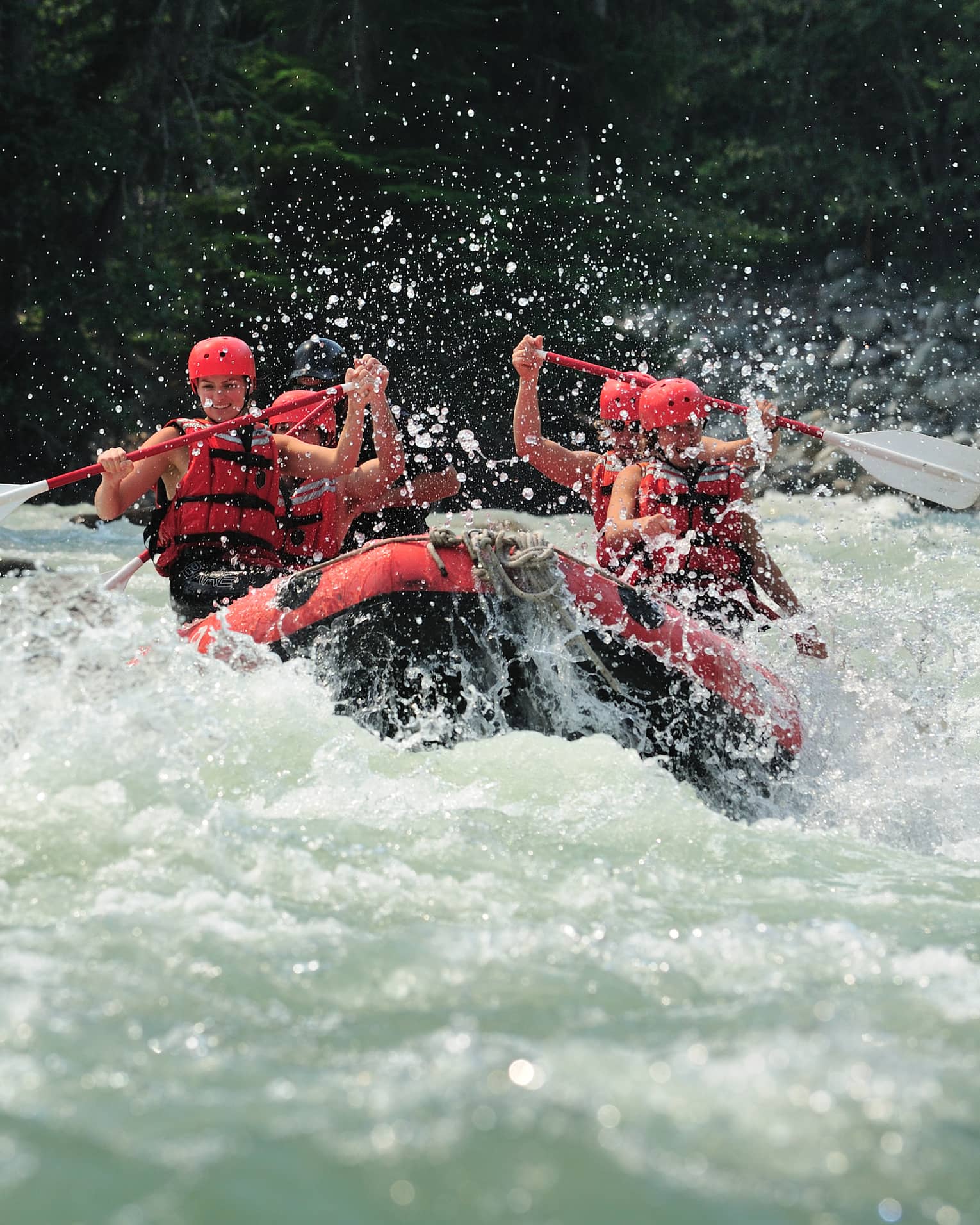 Smiling paddlers in red helmets and lifejackets steer an inflated raft through raging white water, passing evergreen trees.