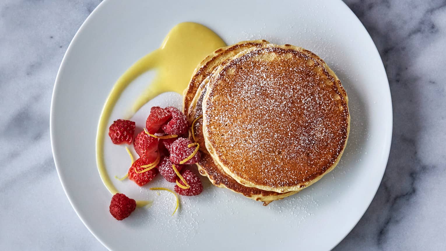 Stack of Lemon Ricotta Pancakes on plate dusted with icing sugar, lemon sauce and fresh raspberry garnish