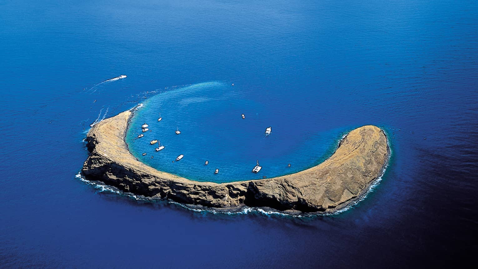 Aerial view of Molokini Crater, a crescent-shaped volcanic islet surrounded by clear blue ocean waters, with boats anchored nearby